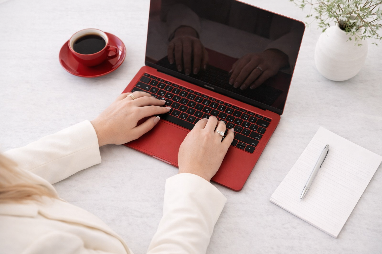 Person typing on a red laptop with a notebook and pen on the white table, a cup of coffee on a red saucer, and a white vase with a green plant.
