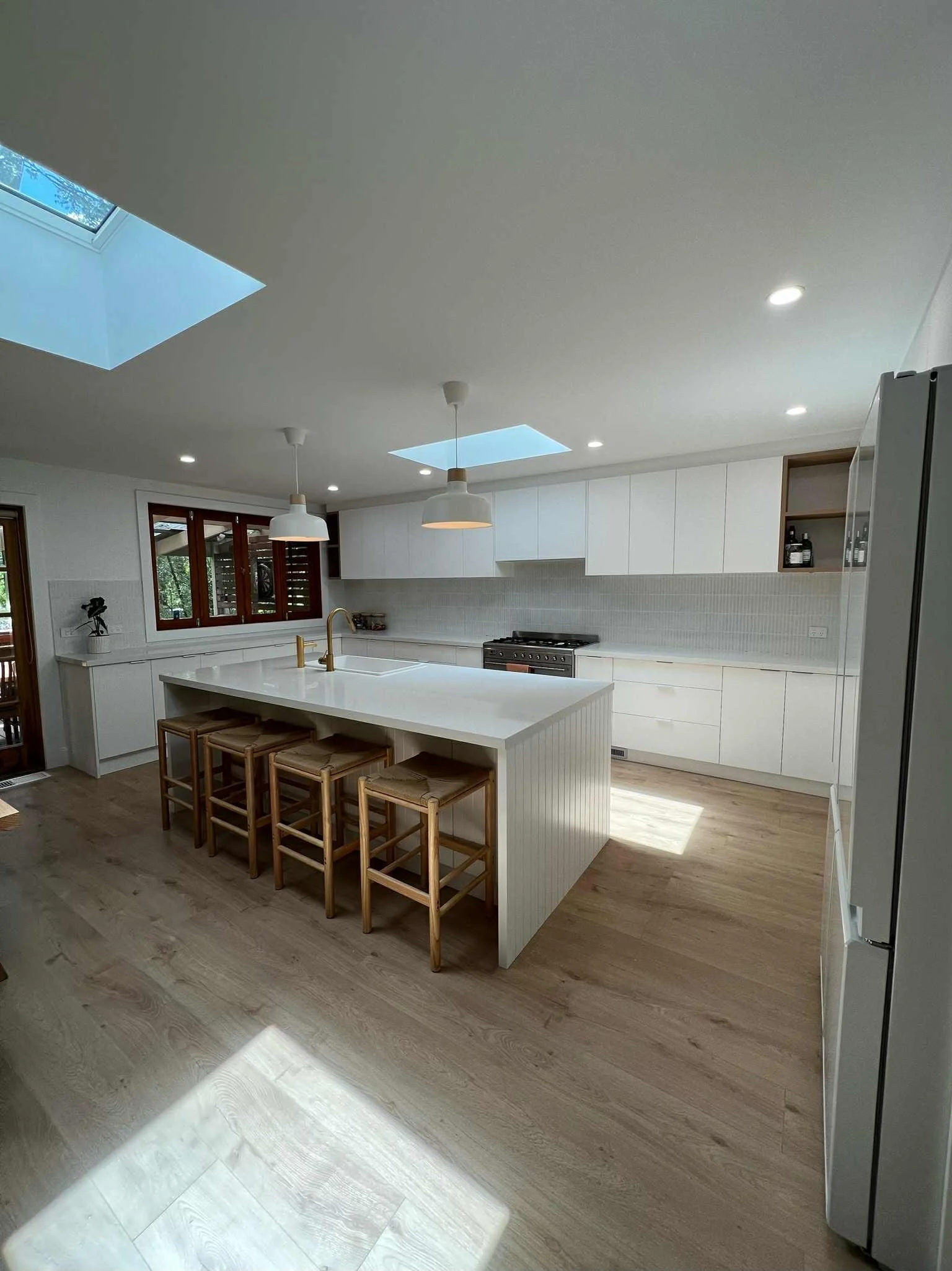 Modern kitchen with white cabinets, large island with seating, wooden stools, stainless steel stove, and two skylights.