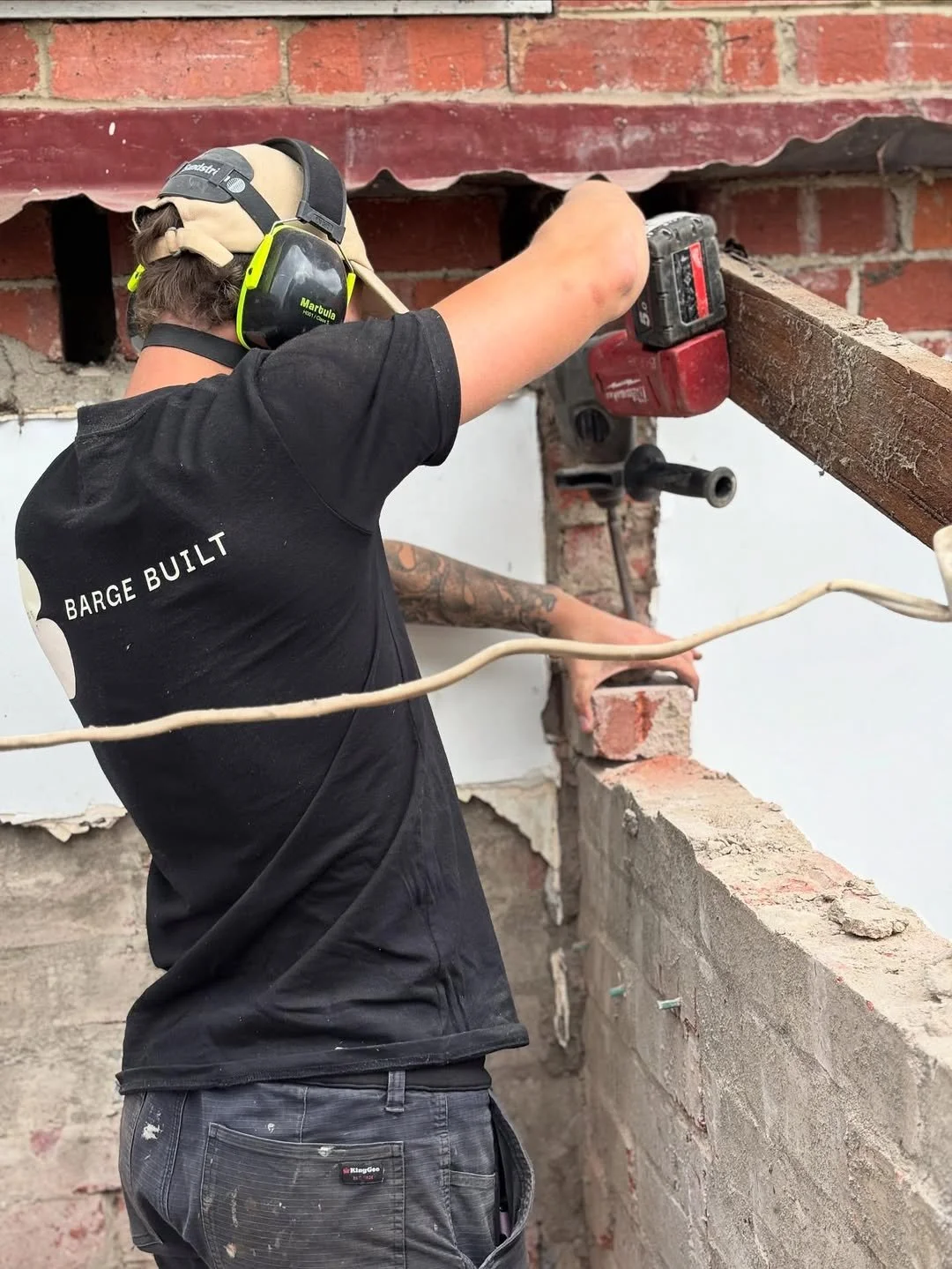 A person wearing a black t-shirt with 'BARGE BUILT' on the back, working with a power drill on a brick wall. The person is wearing safety headphones and has tattoos on their arm.