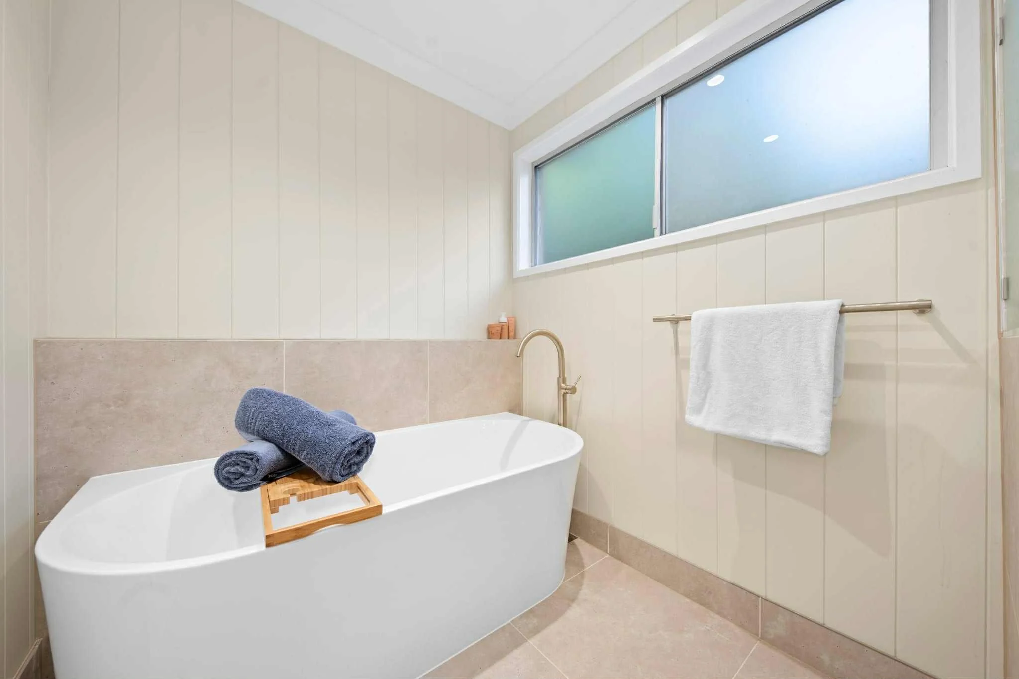 Modern bathroom with free-standing bathtub, two rolled towels on a wooden tray, window, and towel rack with hanging white towel.