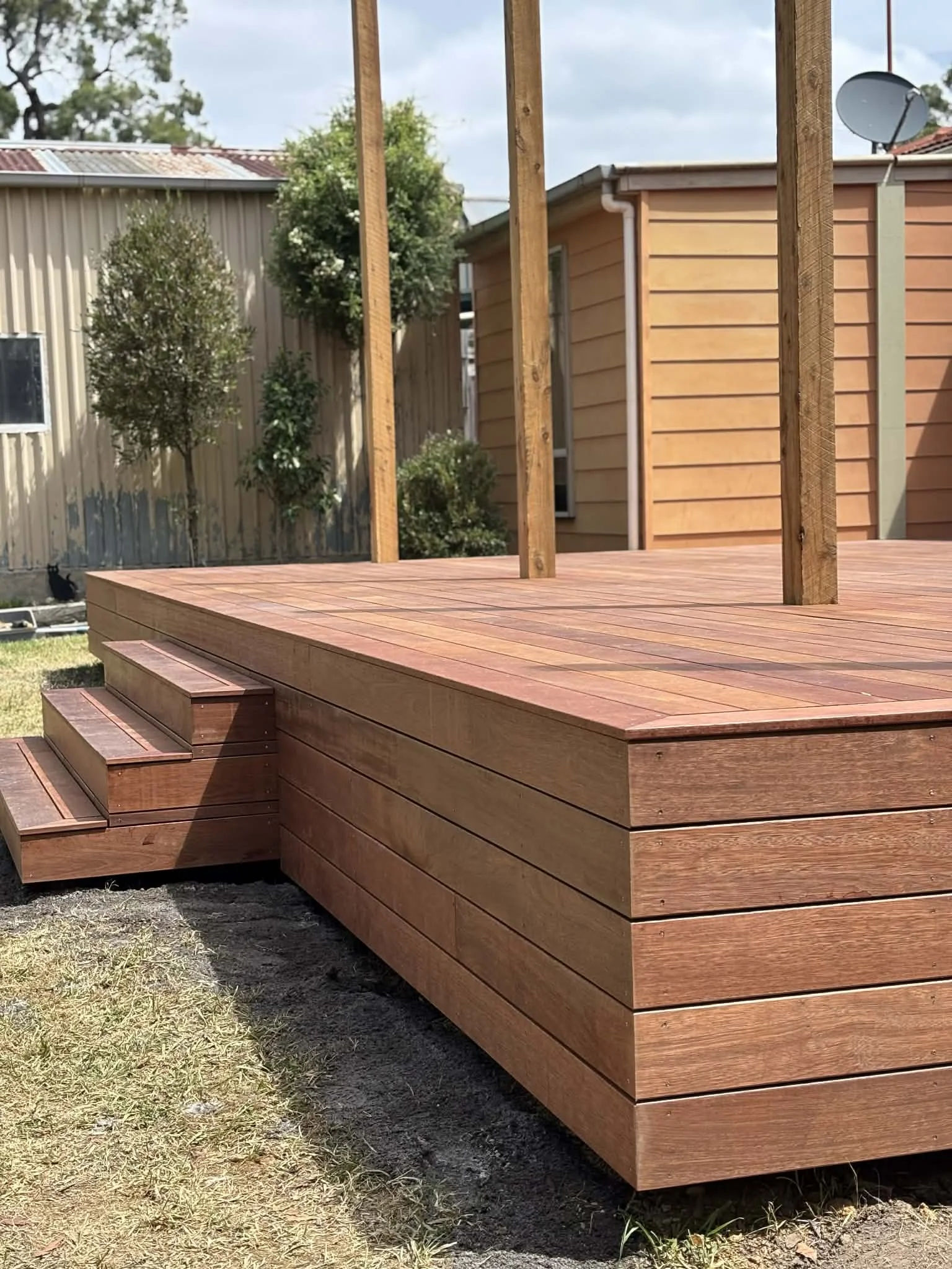 Wooden deck under construction with stairs, in a backyard, with trees, a fence, and garden shed in the background.