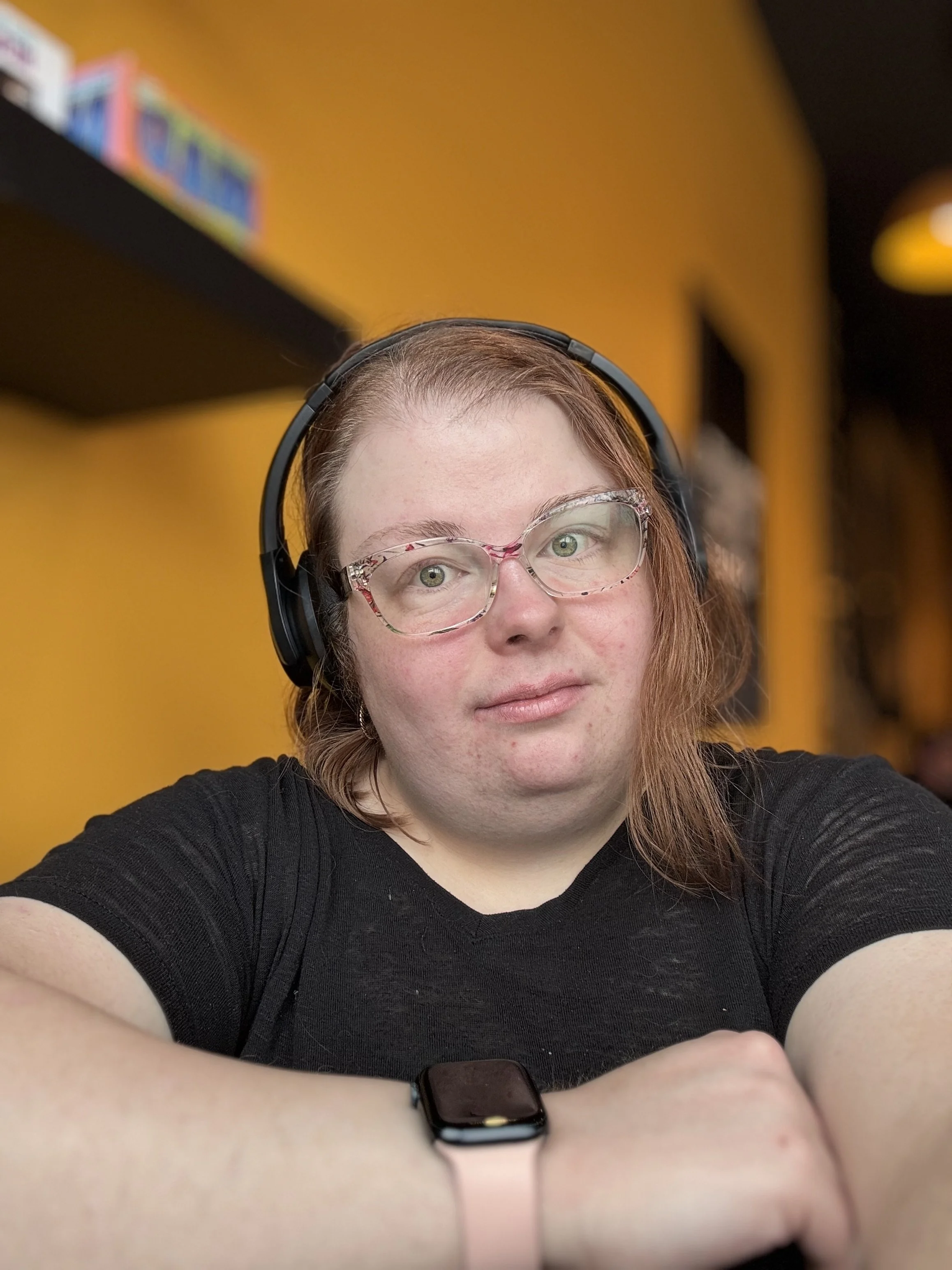 A woman with glasses and headphones looks into the camera, with a yellow wall and bookstore shelf in the background.