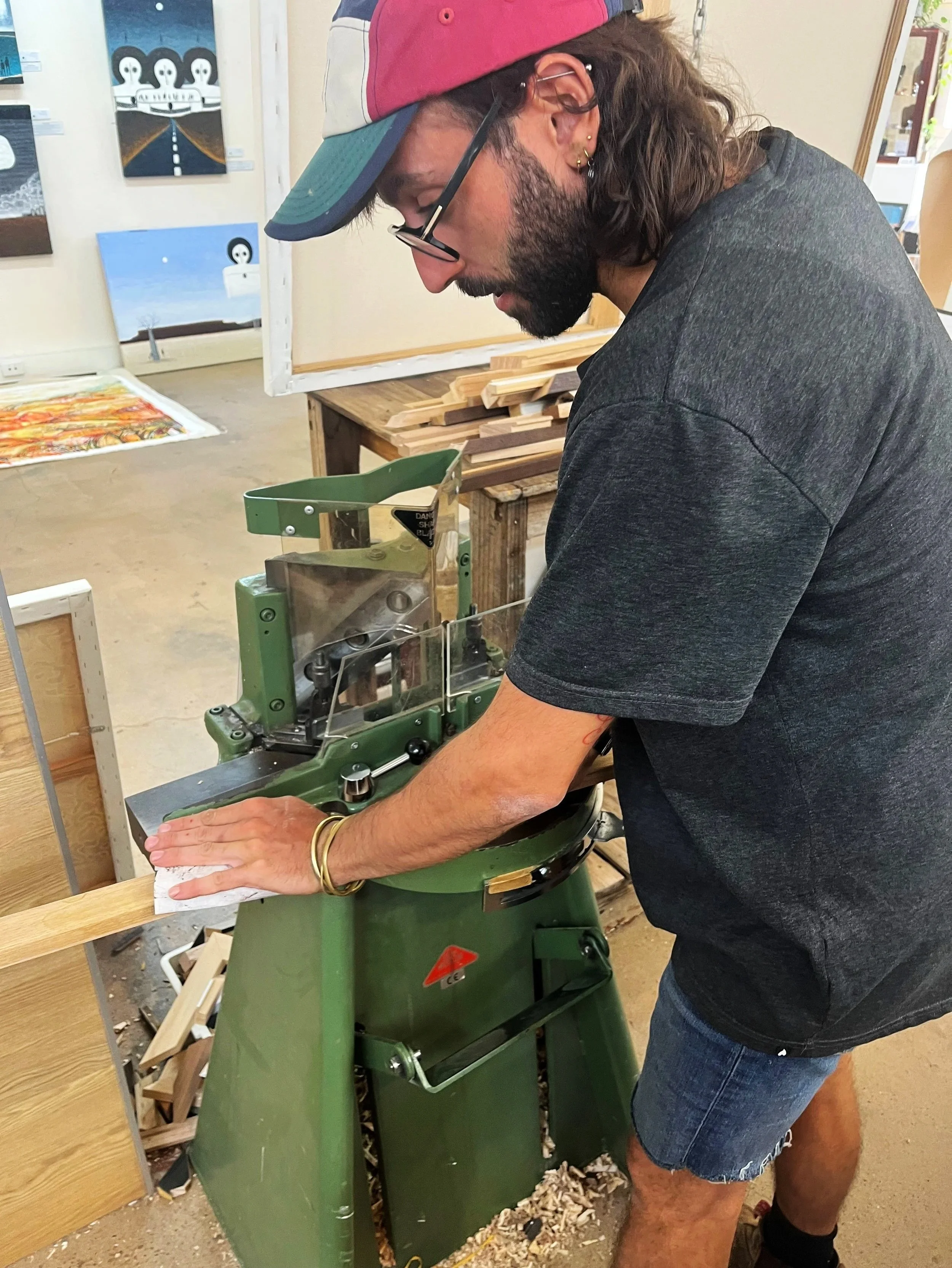 A man with long hair and a beard, wearing a pink and gray cap and glasses, is operating a green woodworking machine in a workshop. He is wearing a dark T-shirt and denim shorts.