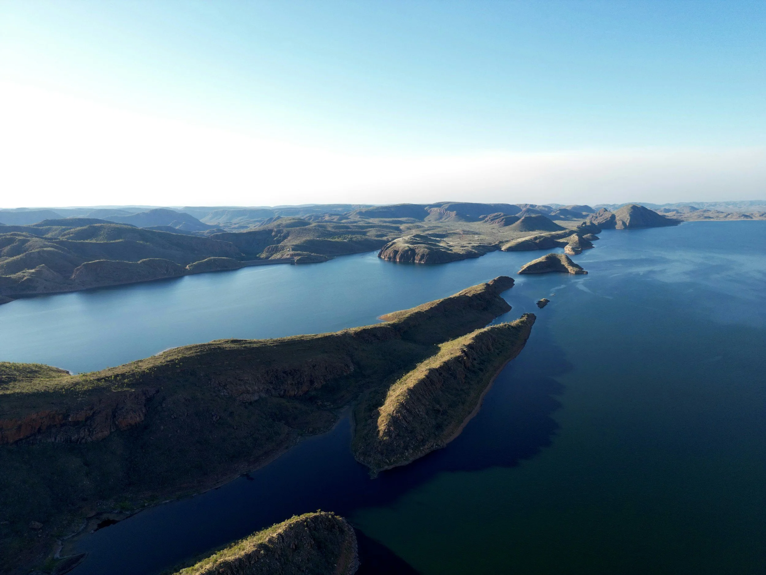 Aerial view of a large body of water surrounded by rolling green hills and small mountains, under a clear blue sky.