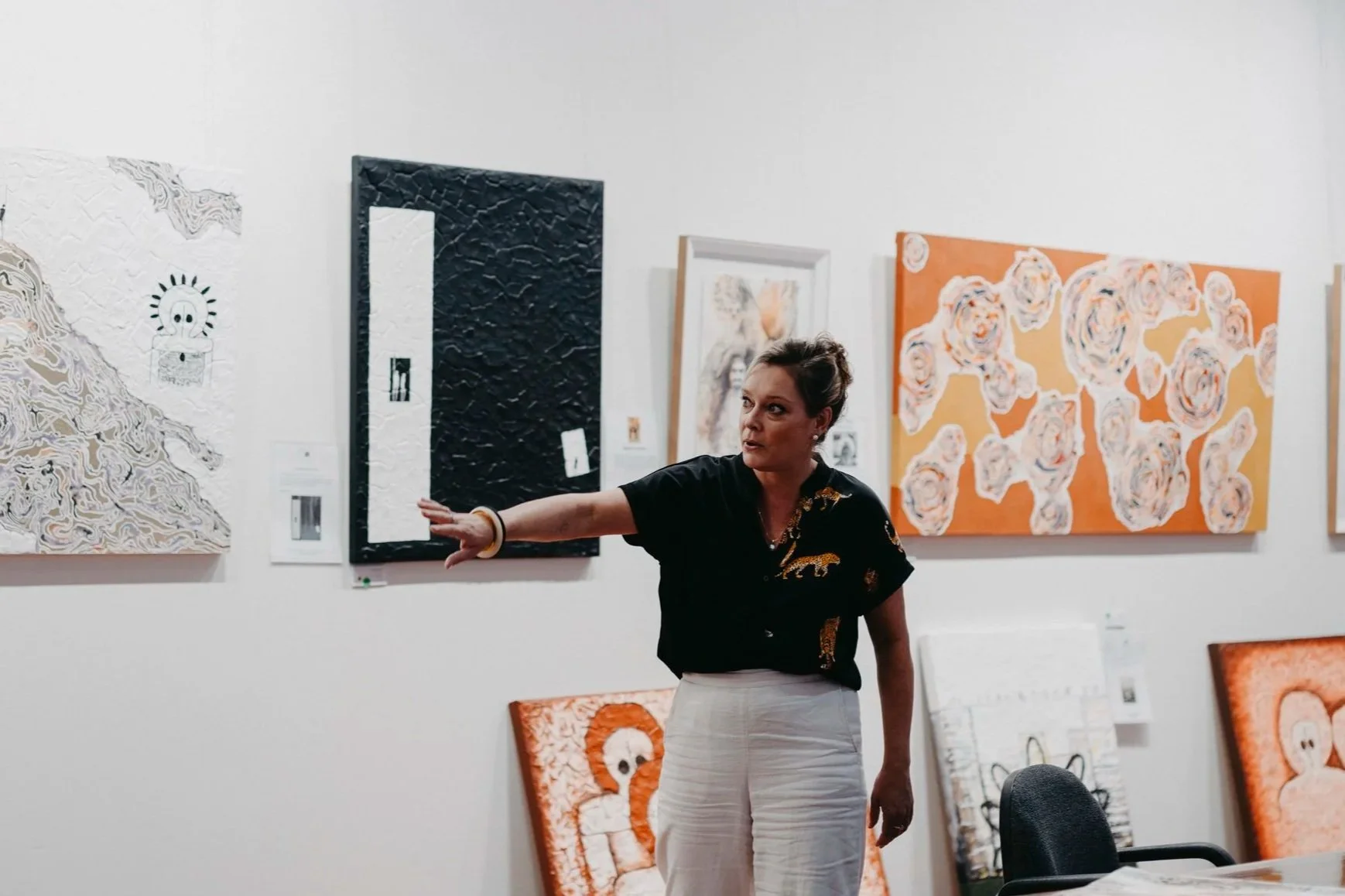 A woman explains artwork in an art gallery, gesturing towards a painting on the wall with her right arm extended, surrounded by various colorful abstract paintings.