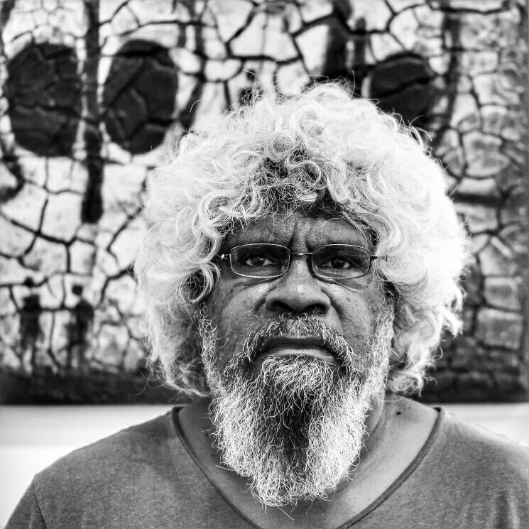 A black and white photograph of a man with voluminous curly white hair, glasses, and a full beard, standing in front of a textured abstract background.