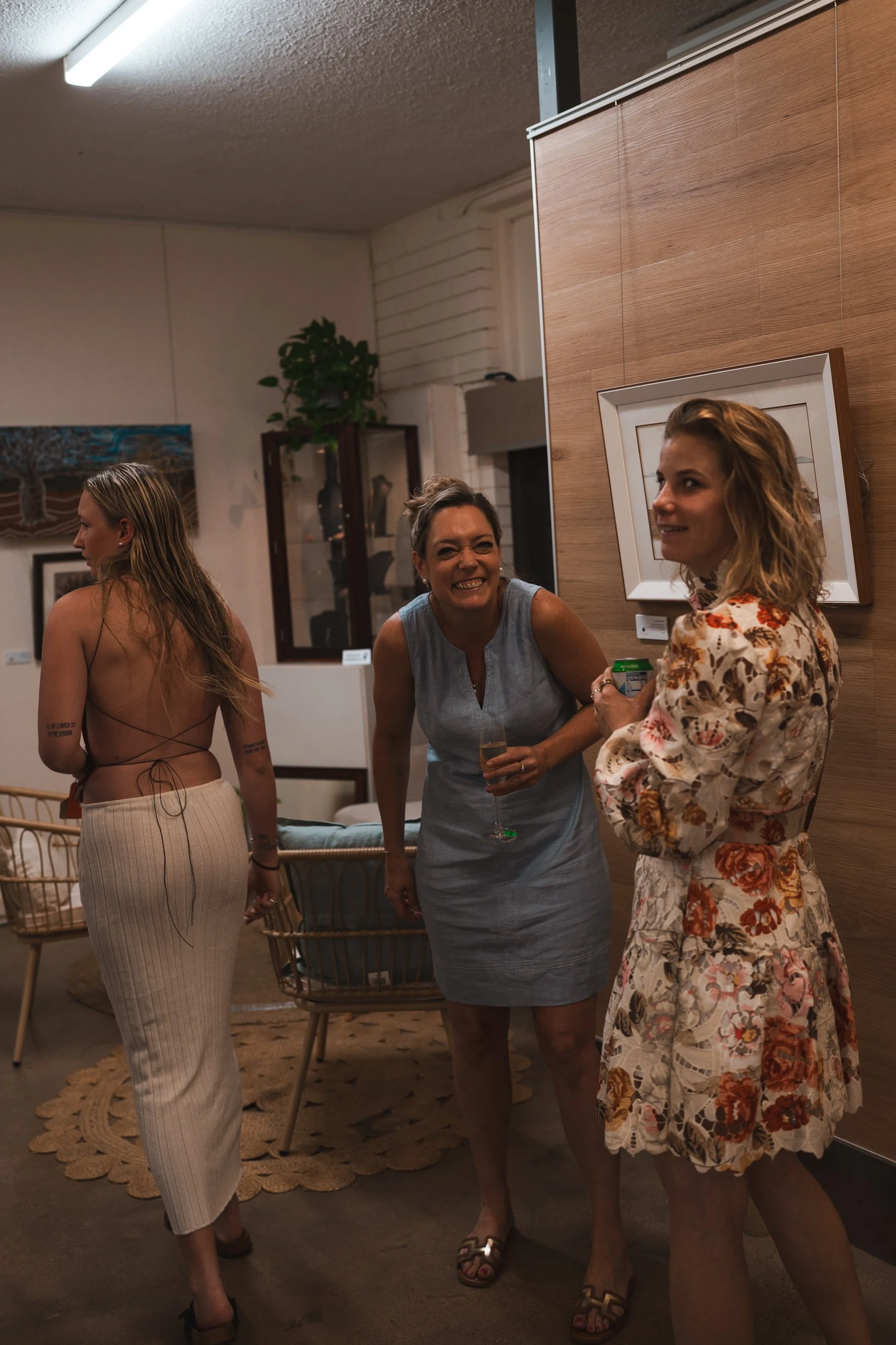 Three women in a social setting, one smiling and two looking away, standing in a room with art on the walls and a wicker table.