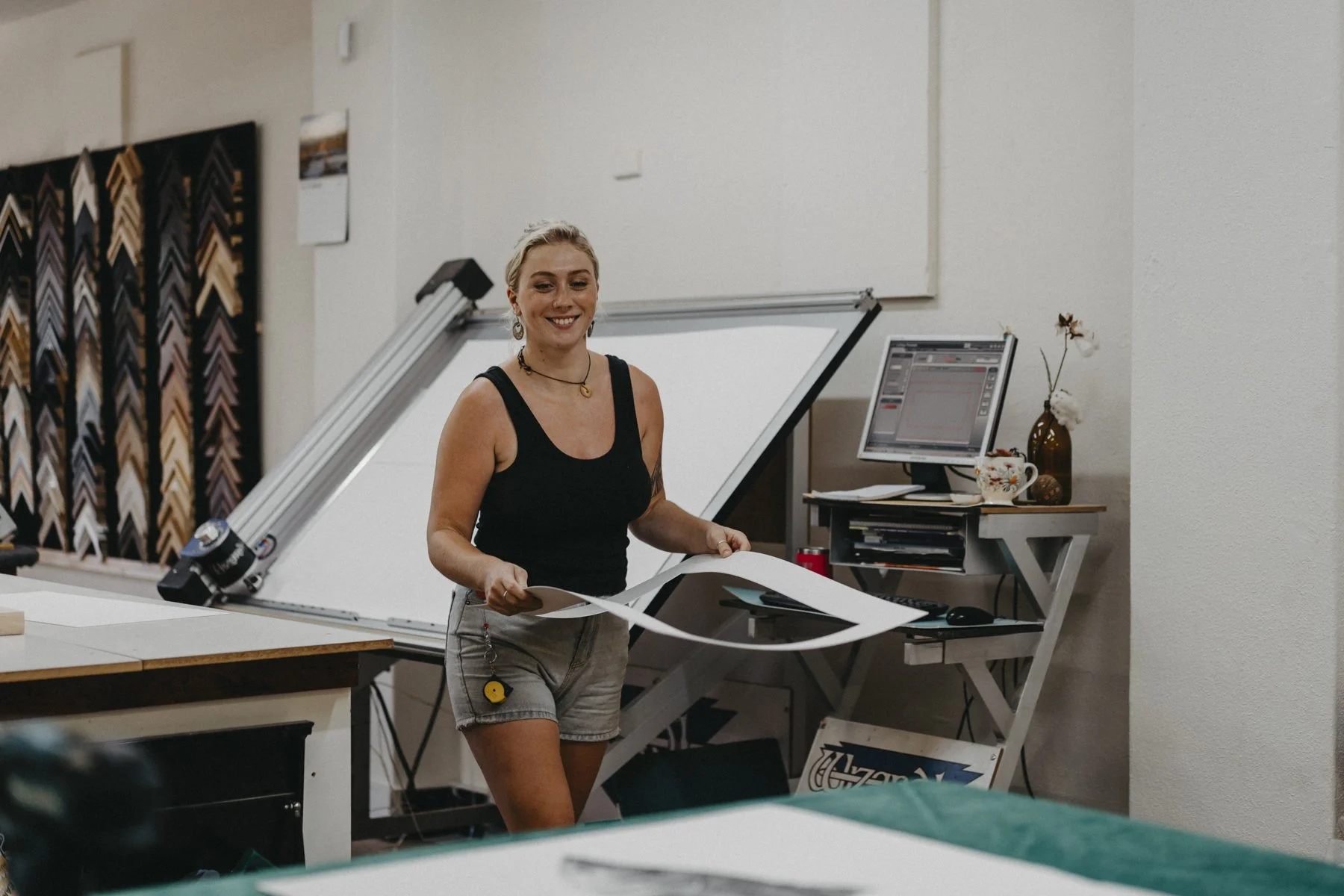 A woman with blonde hair, wearing a black tank top and shorts, is standing in a workspace, holding a sheet of paper and smiling. The workspace contains a drafting table, a computer with a monitor, and various art supplies, with framed samples of artwork on the wall.