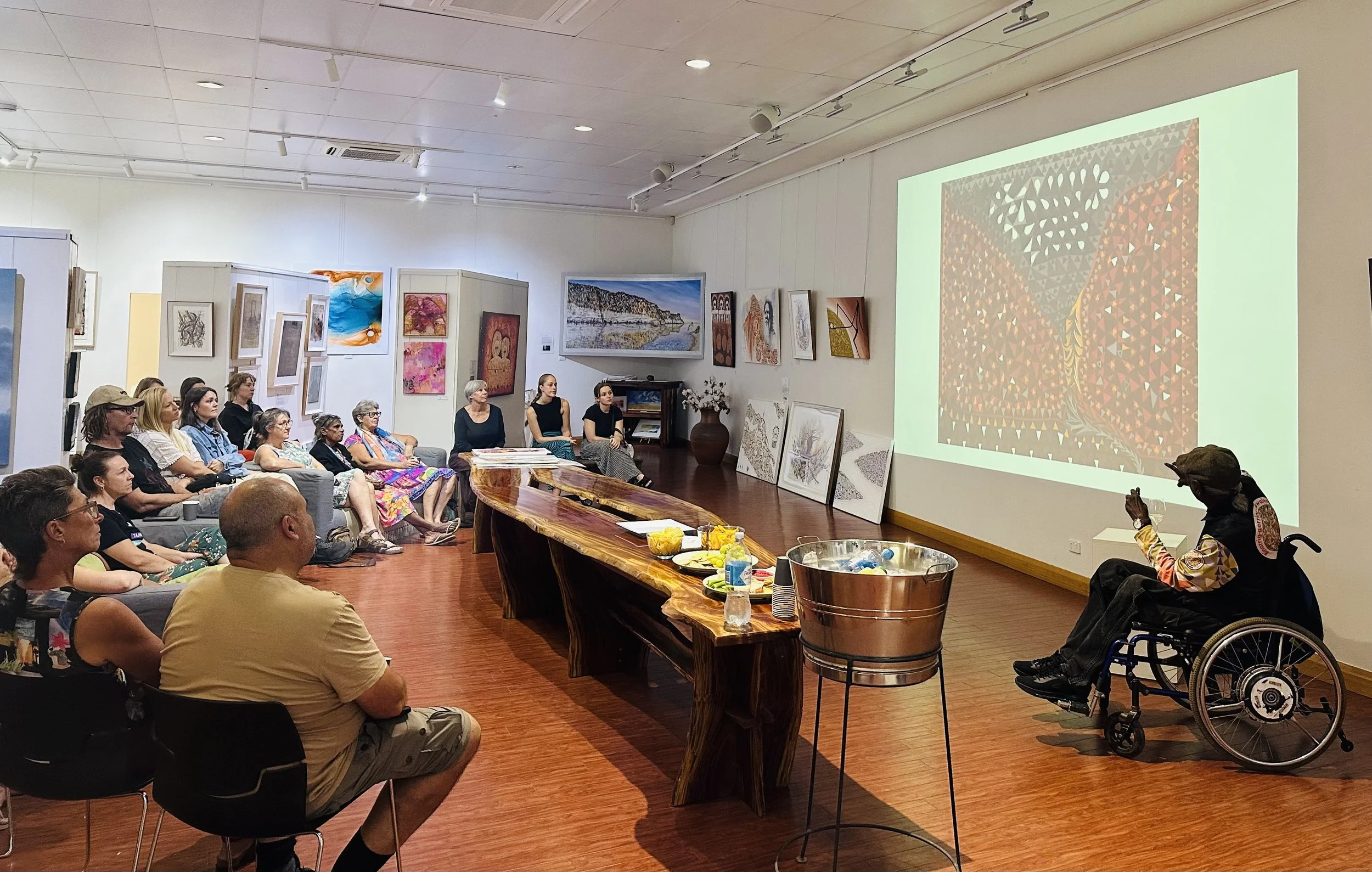 An art gallery event with a diverse group of people seated and watching a presentation on a large screen displaying a colorful abstract artwork. Art pieces are displayed on the walls, and a table with snacks and drinks is visible at the front.