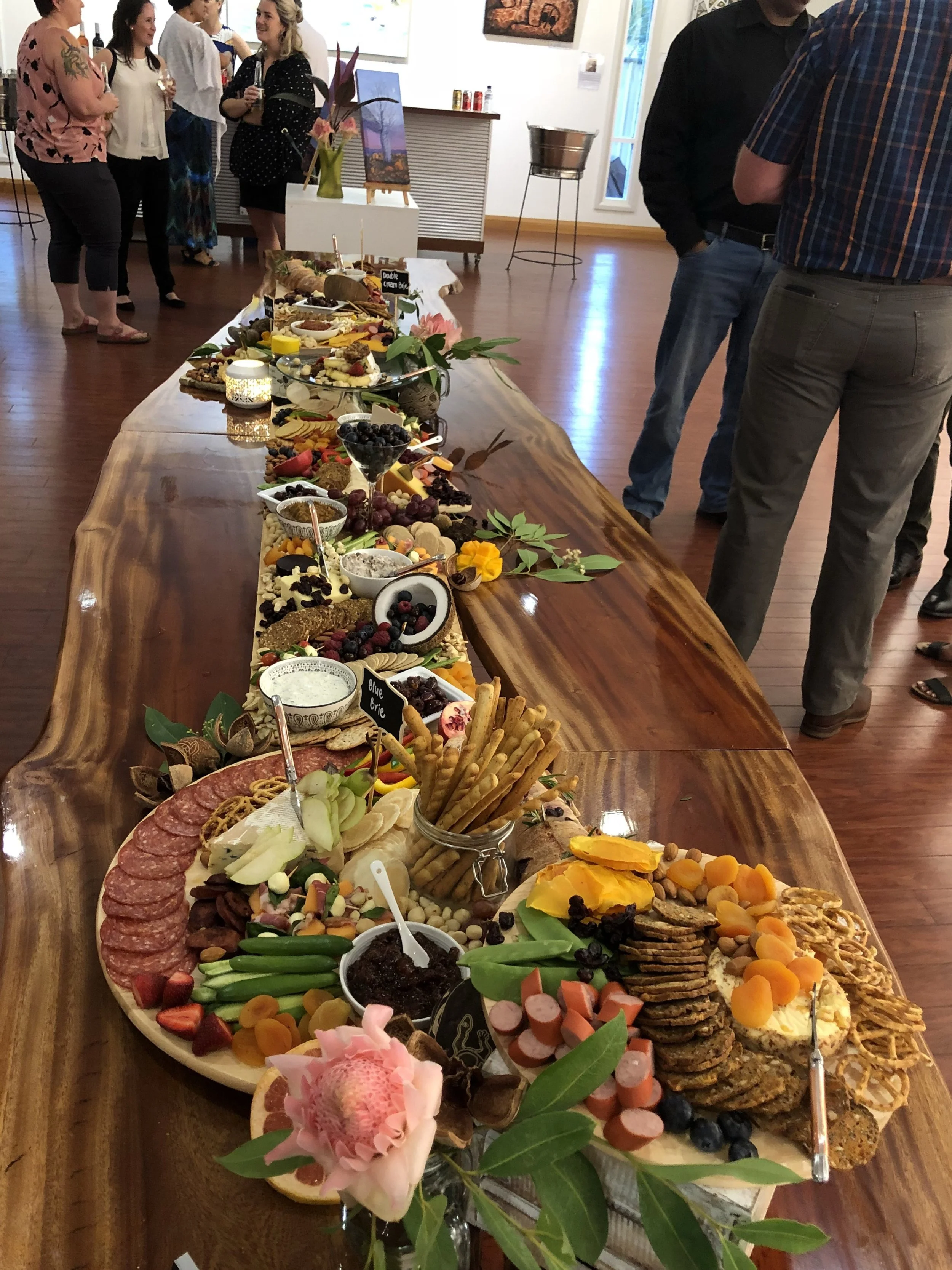 A long wooden table filled with various cheeses, fruits, crackers, and spreads, decorated with flowers, at a social gathering with people standing and chatting in the background.