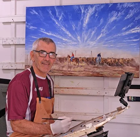 An artist smiling while painting a landscape of sky and cattle on a canvas in a studio.