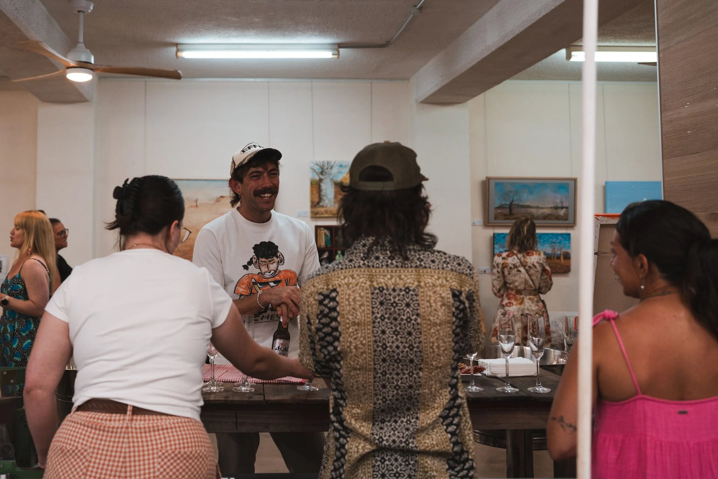 People gathered at a social event, with a man in a white cap smiling and holding drinks, engaging in conversation. The setting appears to be an art gallery or similar venue, with paintings hanging on the wall.