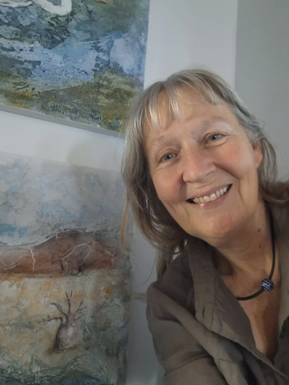 A smiling woman with gray hair standing next to a painting of a tree and a landscape.