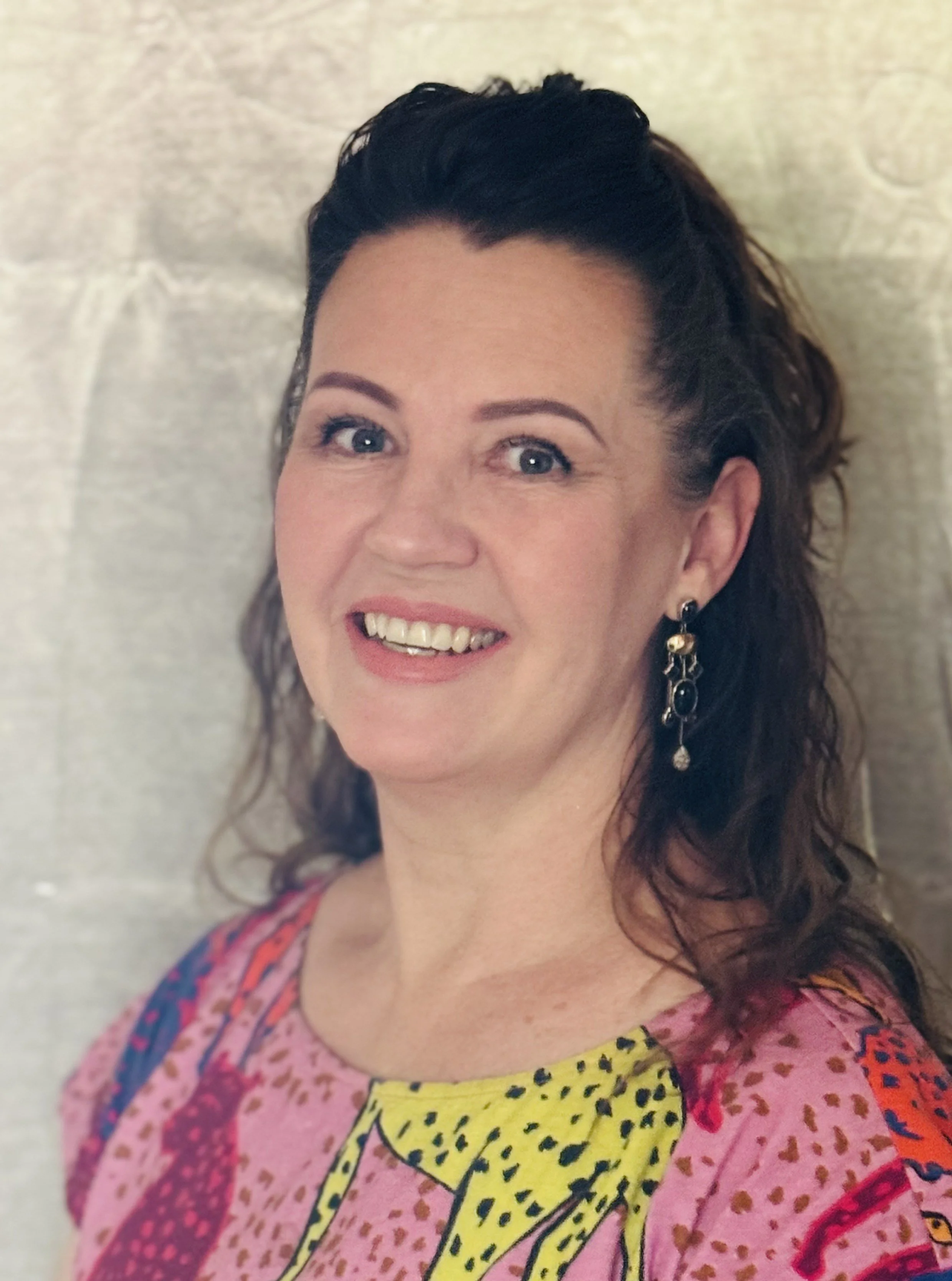 A woman with dark hair smiling, wearing earrings and a colorful patterned dress, standing in front of a textured wall.