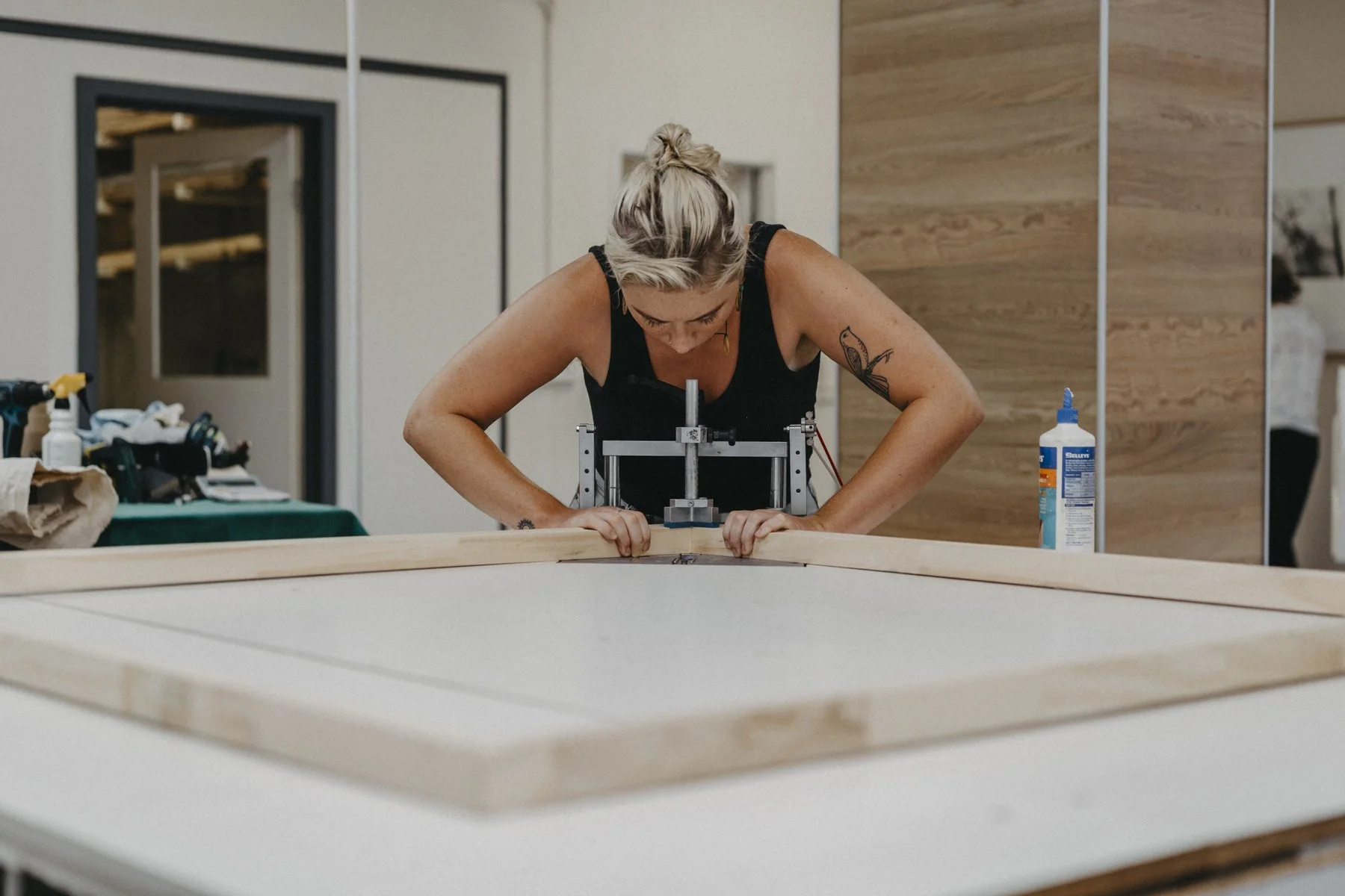 A woman with blonde hair tied up in a bun, working on a woodworking project at a table. She's using a tool to measure or cut wood, with a bottle of glue nearby, in a workshop or craft room.