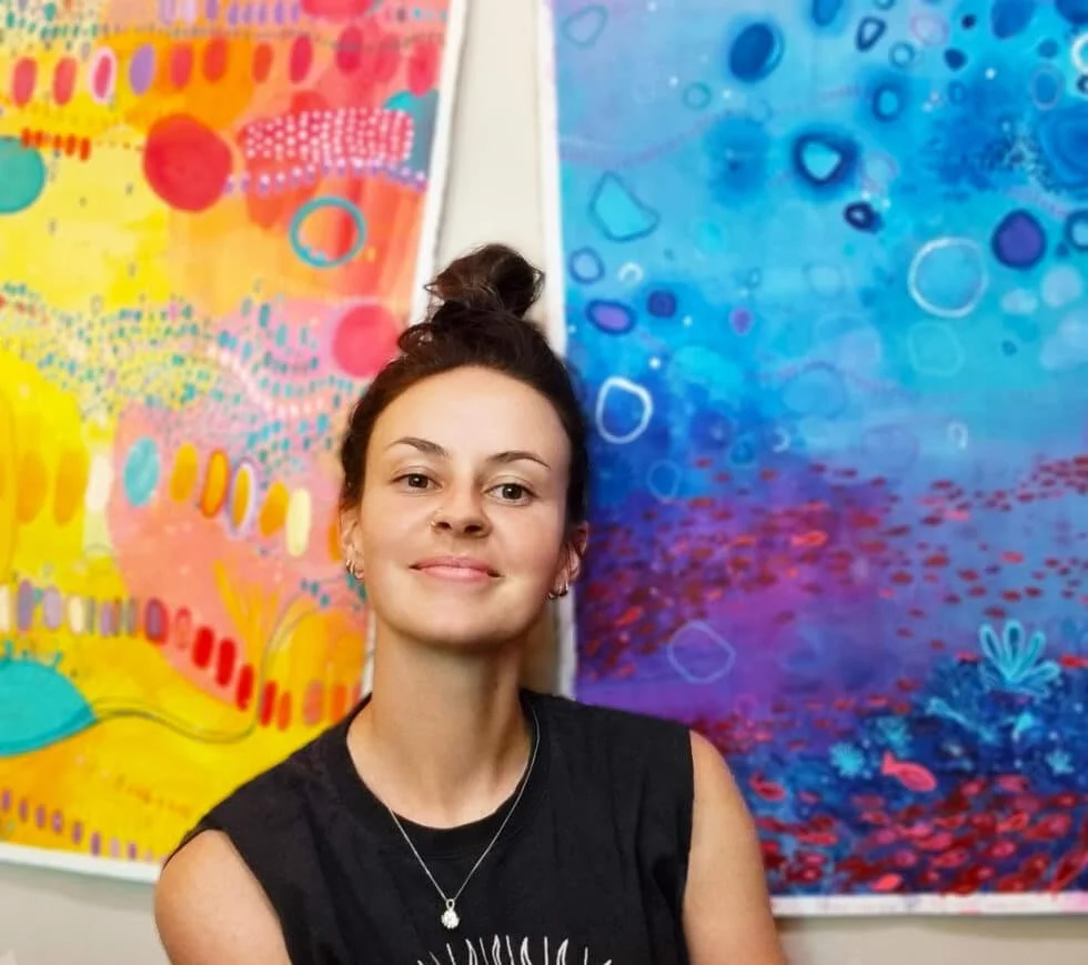 Young woman with dark hair tied in a bun, wearing a black sleeveless shirt, standing in front of colorful abstract paintings with bright yellow, pink, blue, and purple patterns.