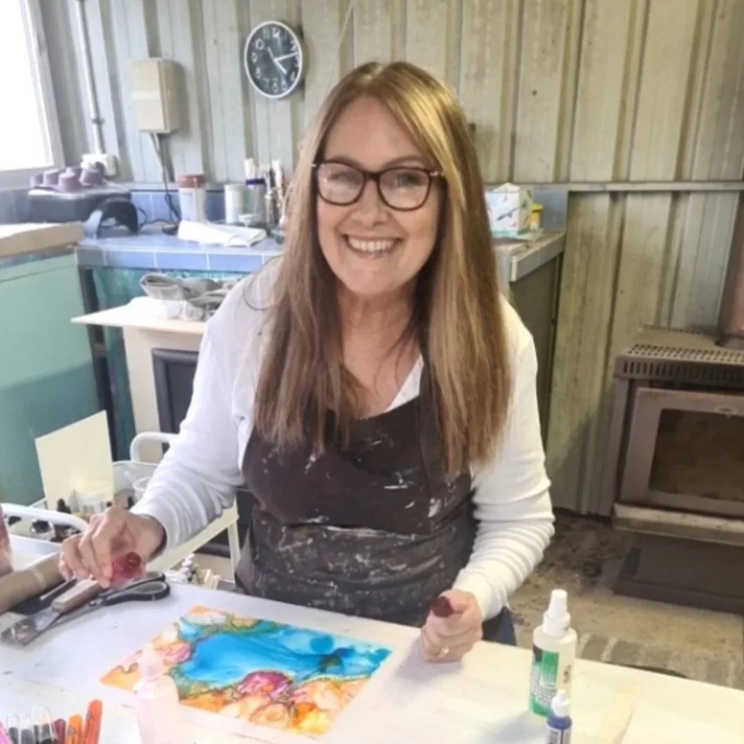 A woman with long red hair and glasses smiling at a craft table with paints and tools, in a rustic kitchen setting.