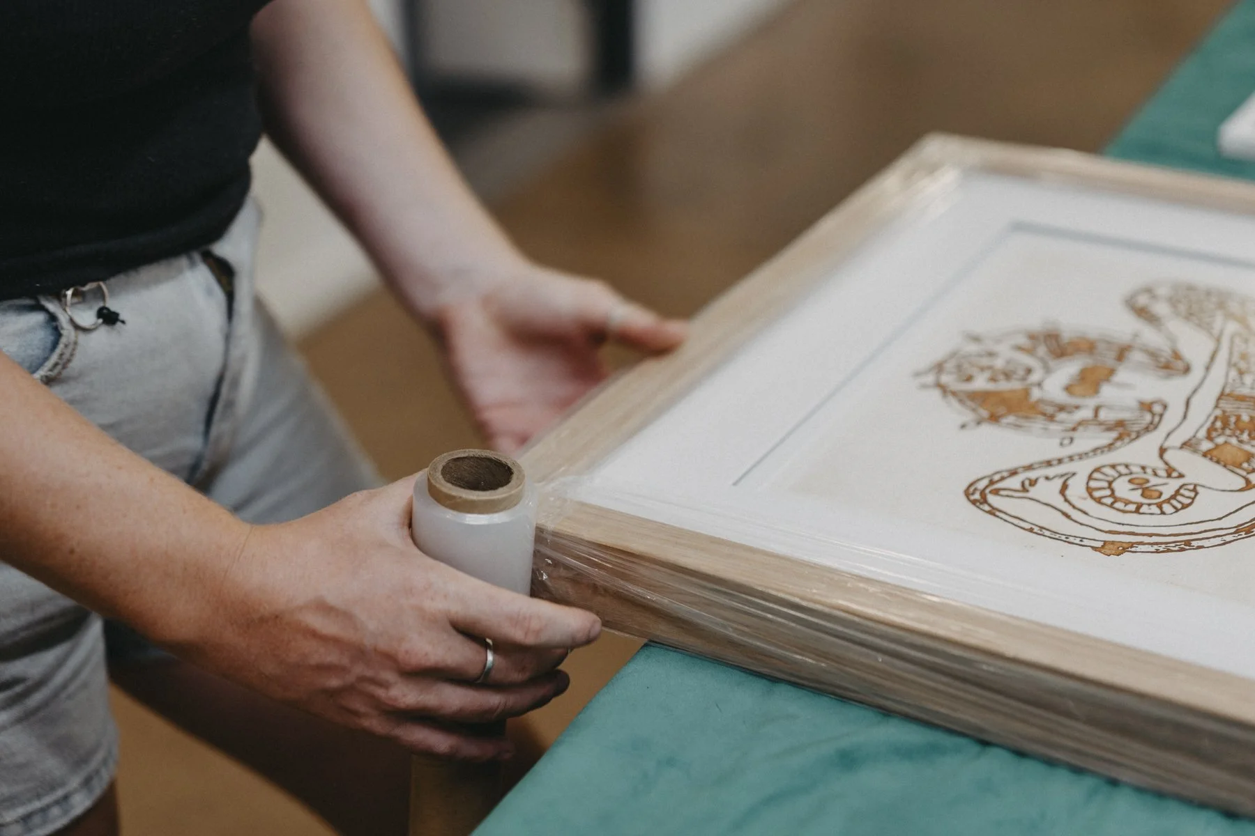 Person applying a protective plastic wrap to a framed artwork on a table.