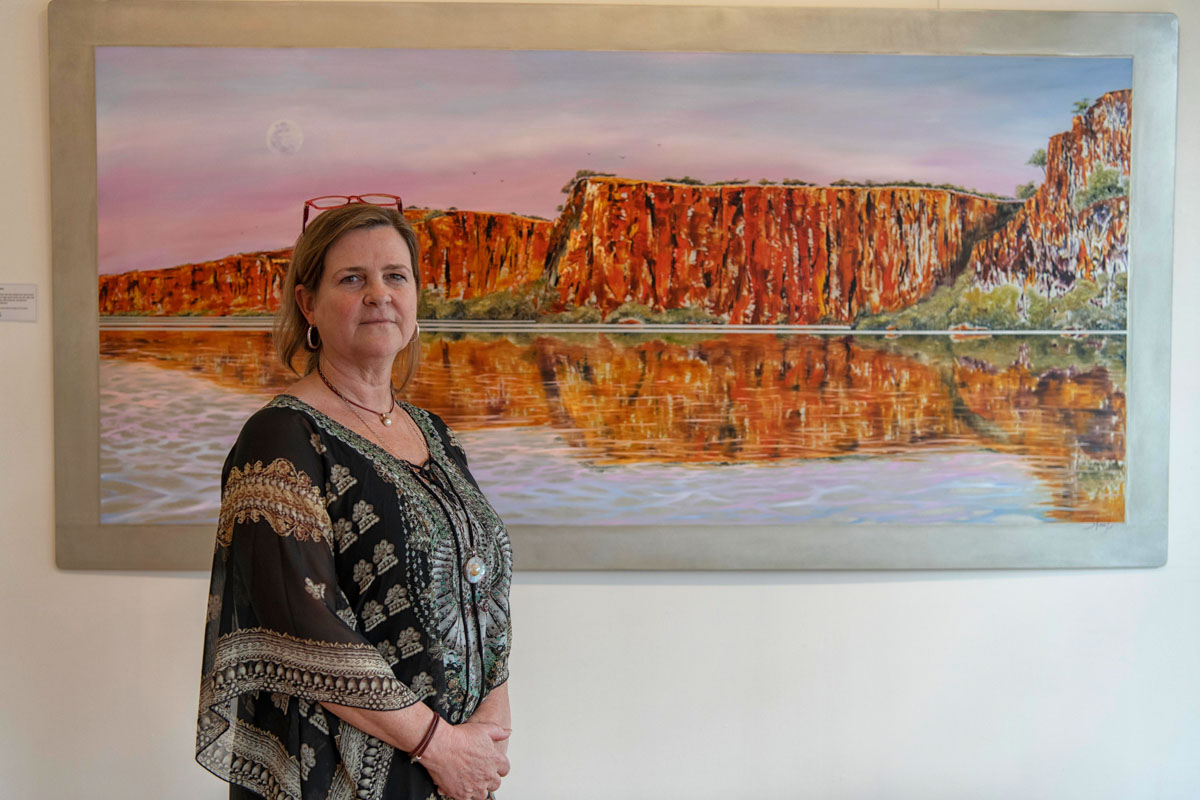 A woman standing in front of a large painting of a river with orange cliffs and a purple sunset sky.