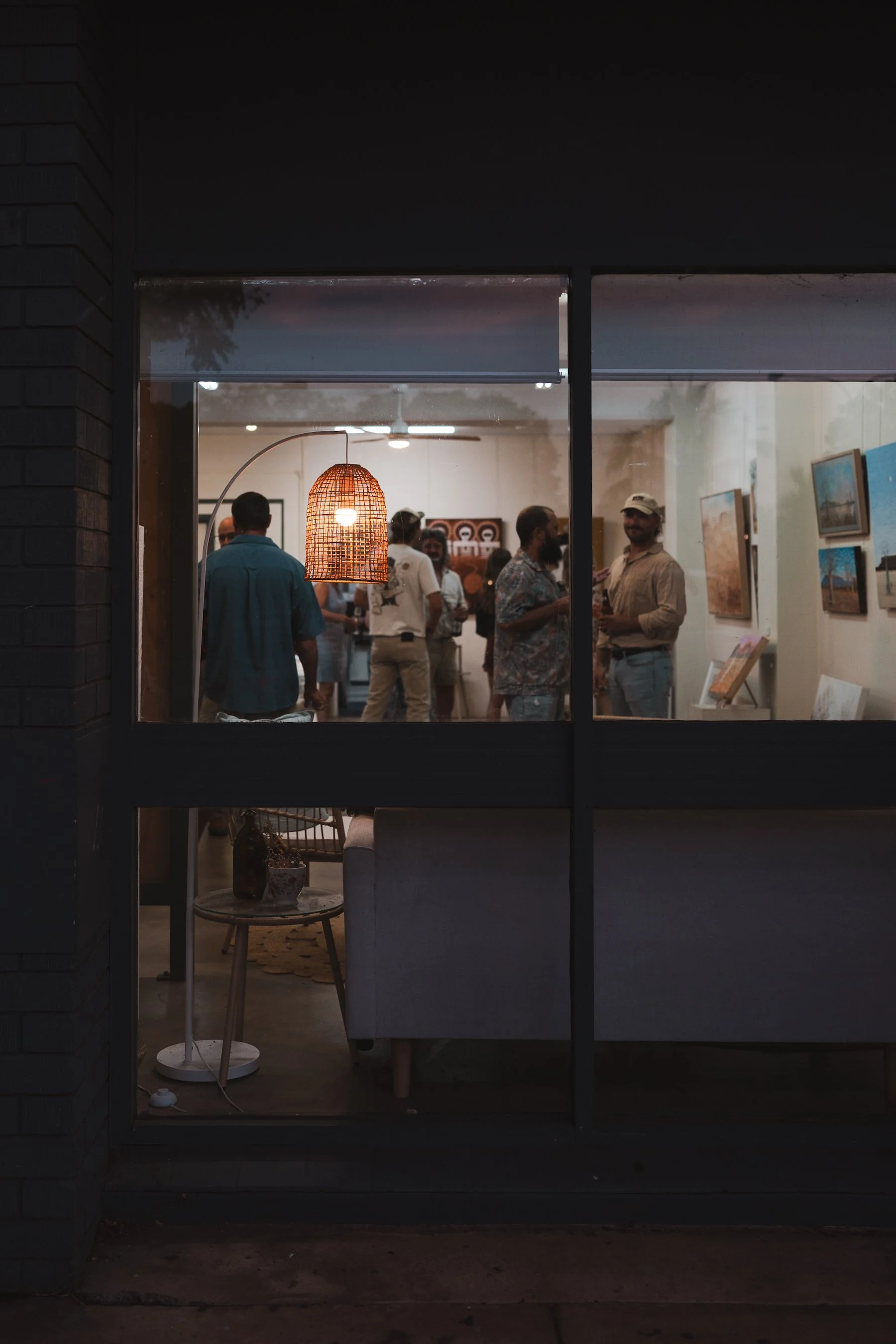 View through a window into an art gallery with several people socializing. There is a rattan pendant light hanging from the ceiling, and paintings on the walls.