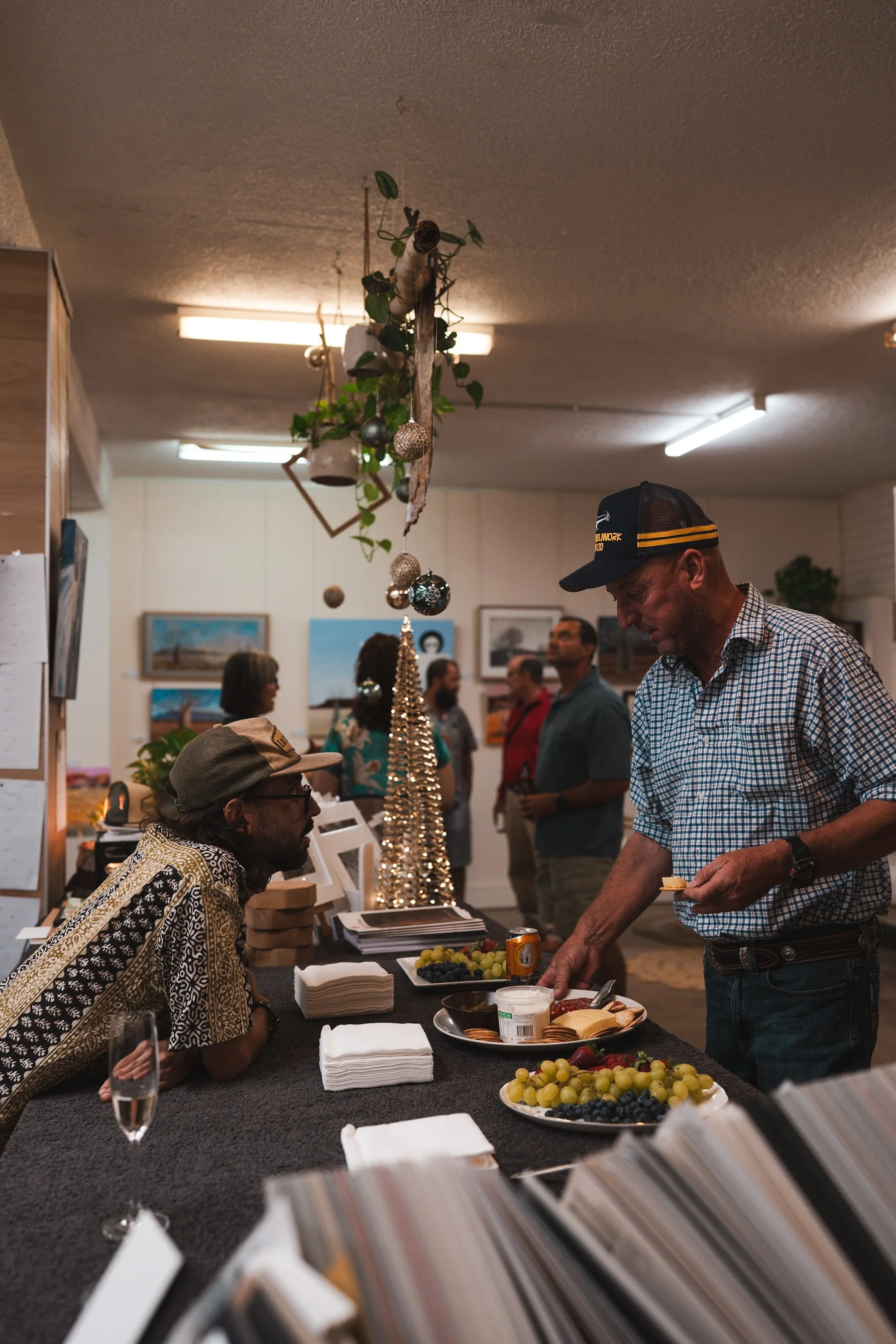 People at a table or stand with fruit and snacks, indoors with hanging plants and artwork, some guests talking and browsing, and a man serving himself
