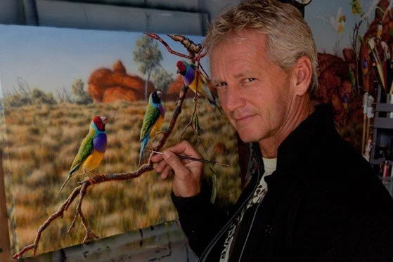 A male artist painting a desert landscape with three colorful parrots perched on a branch in the foreground.