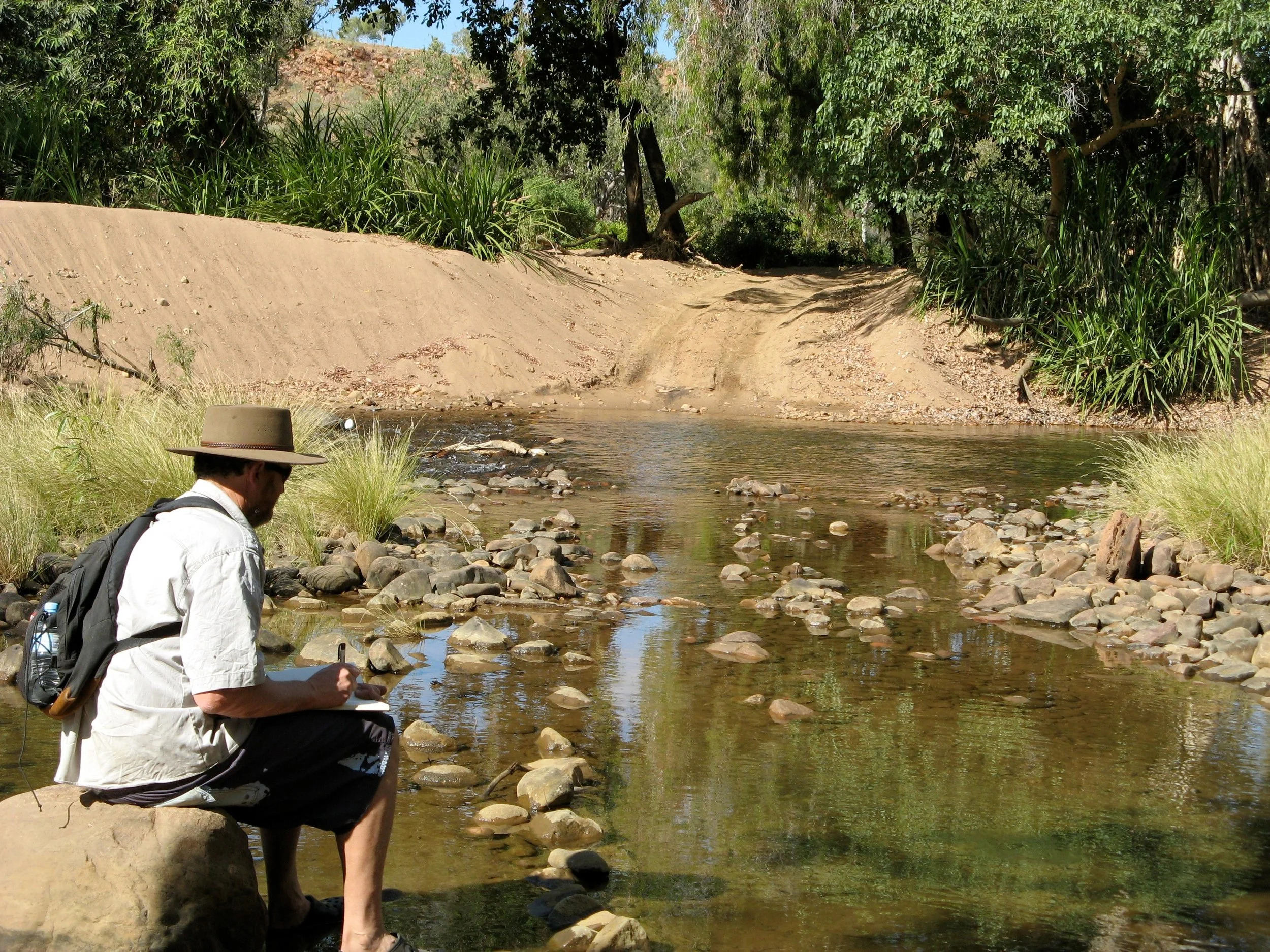 A person wearing a hat and white shirt sitting on a rock by a river, surrounded by green trees and plants, taking notes or drawing.