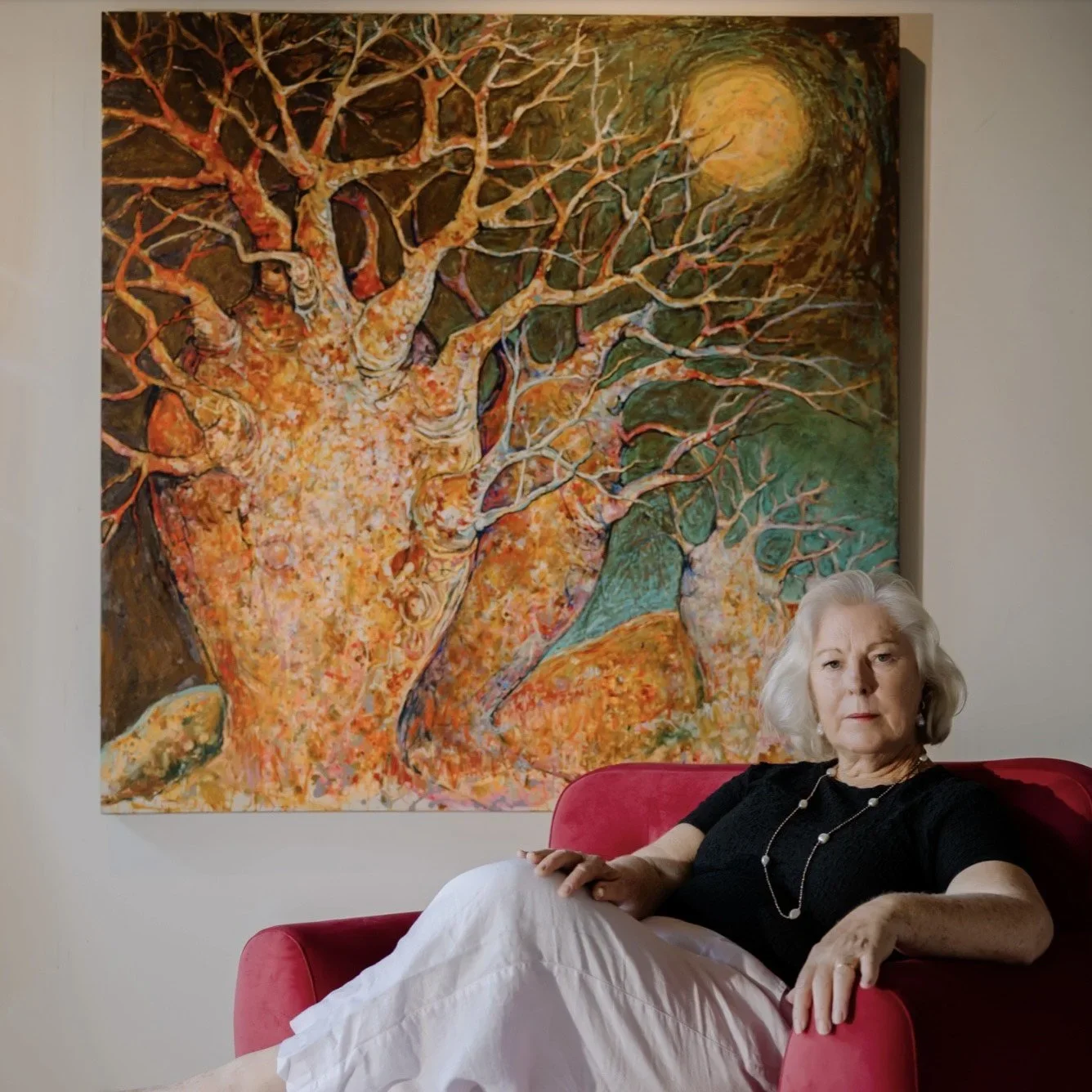 An elderly woman with white hair sitting on a red sofa in front of a large colorful abstract painting of a tree with many branches and a bright sun or moon in the background.