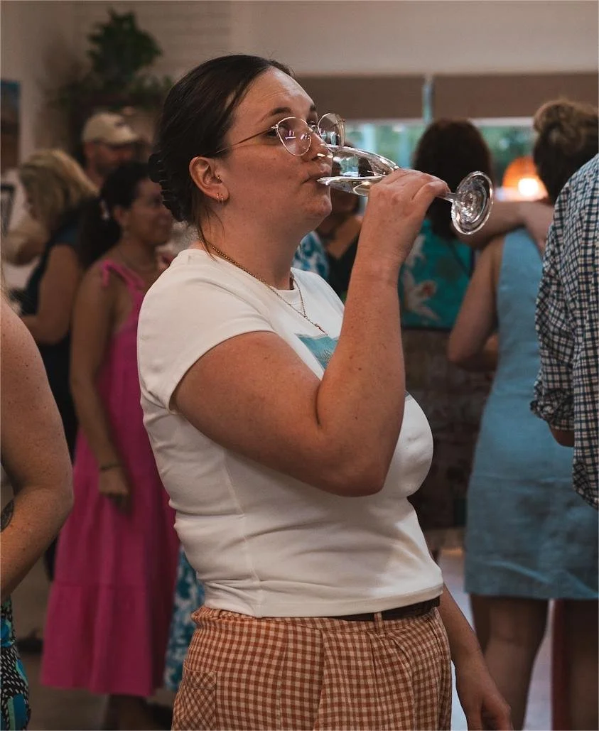 A woman with glasses and dark hair, wearing a white T-shirt and checkered orange pants, drinking from a wine glass at a social gathering indoors.