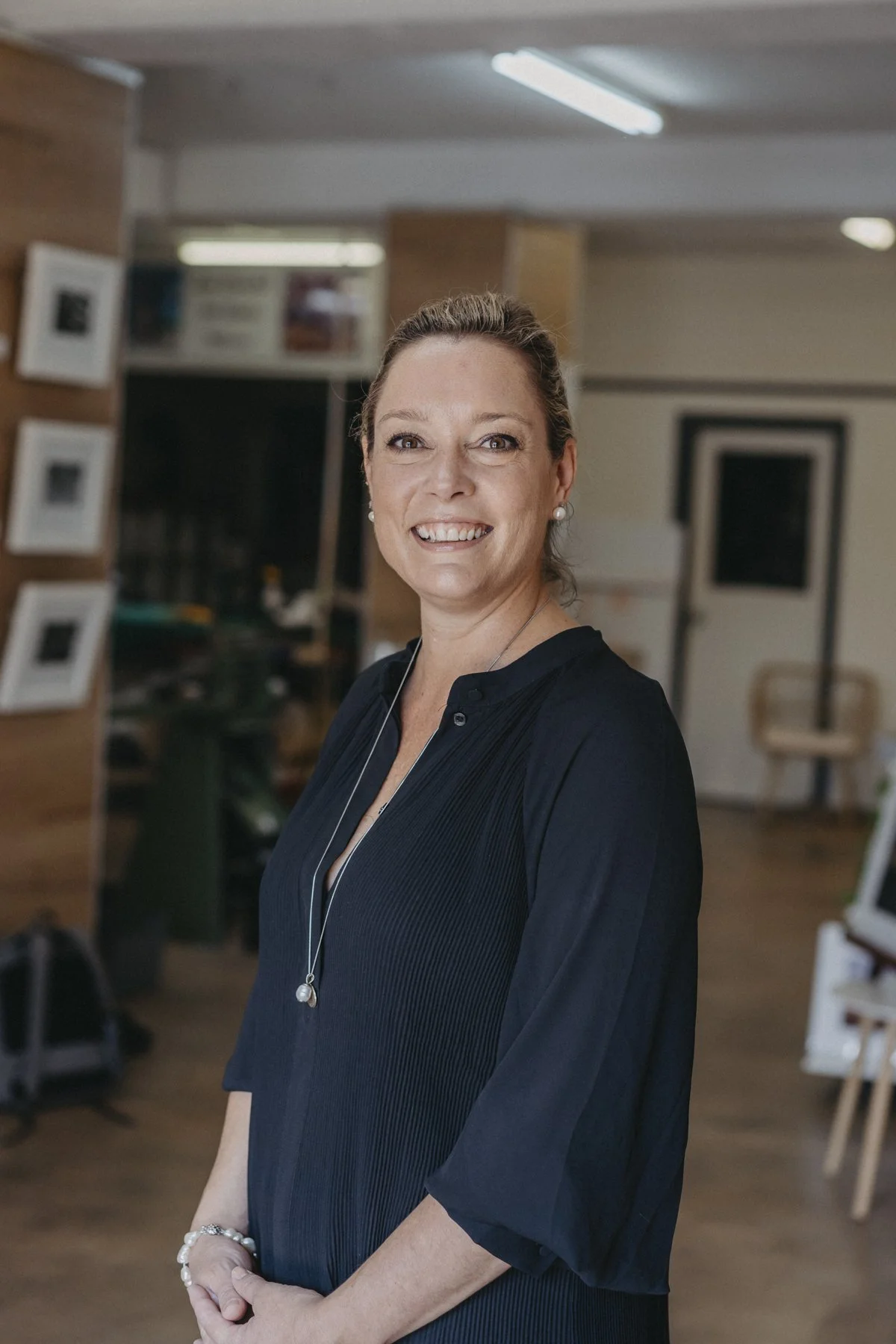 Portrait of a smiling woman with brown hair, wearing a black dress and jewelry, standing indoors in a room with picture frames on the wall and soft lighting.