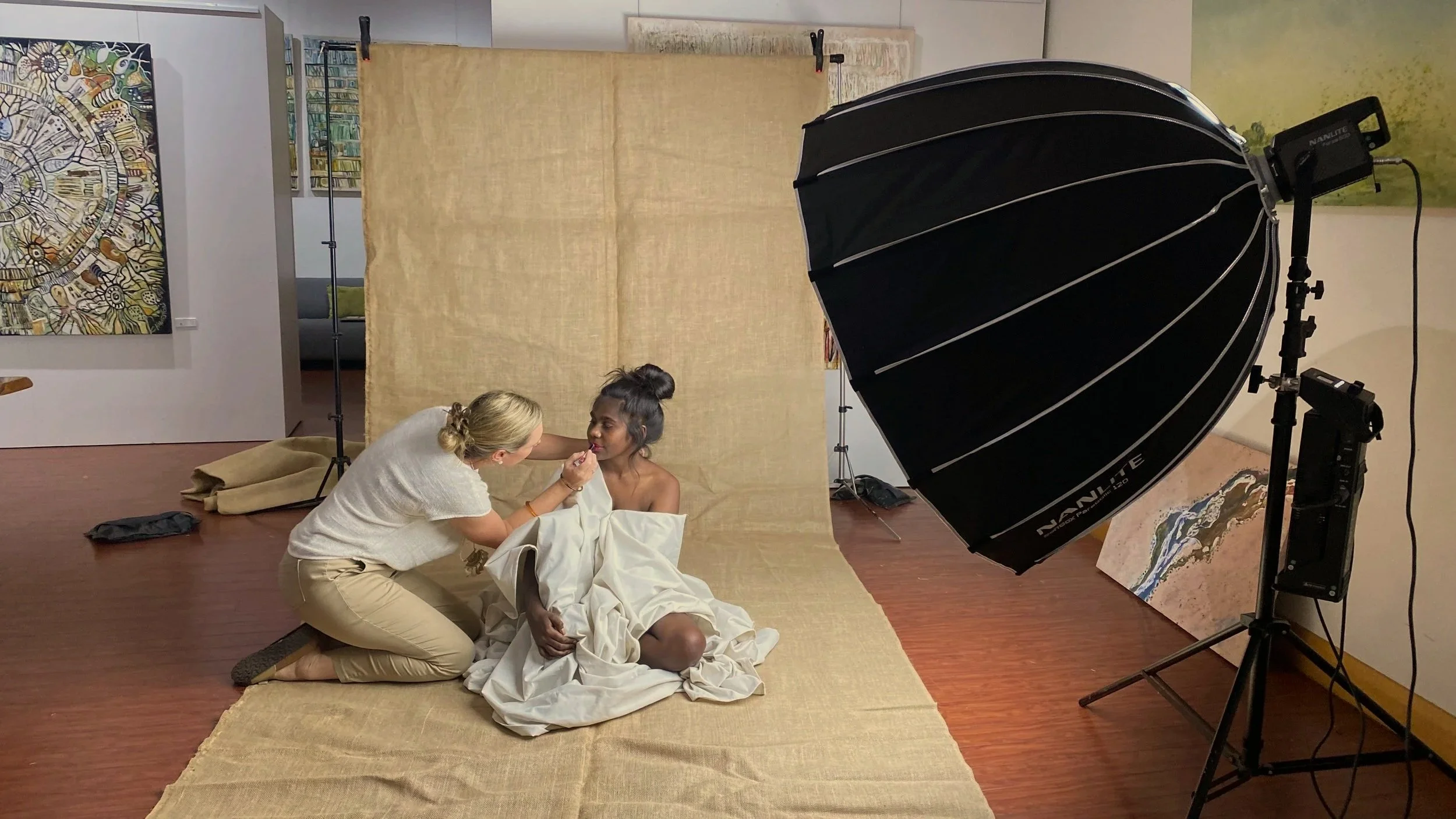 Photographer applying makeup or doing touch-up on a model sitting on the floor in an art studio with paintings and beige fabric backdrop.