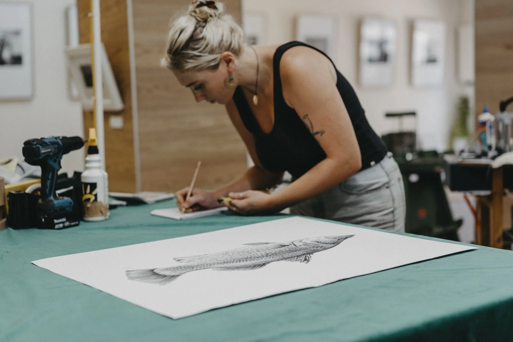 A woman with short blonde hair wearing a black tank top and beige pants is drawing or writing on a notebook at a workspace. On the table in front of her, there is a detailed black and white fish drawing, a cordless drill, and various supplies. The background features a wooden wall and framed pictures.