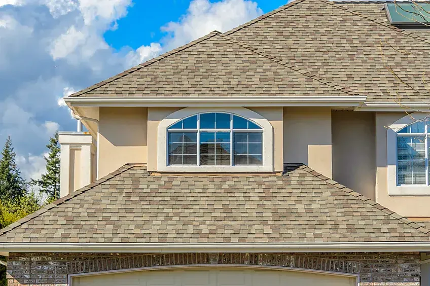 Close-up of the upper section of a house with beige walls, a shingled roof, and large arched windows on the second floor.