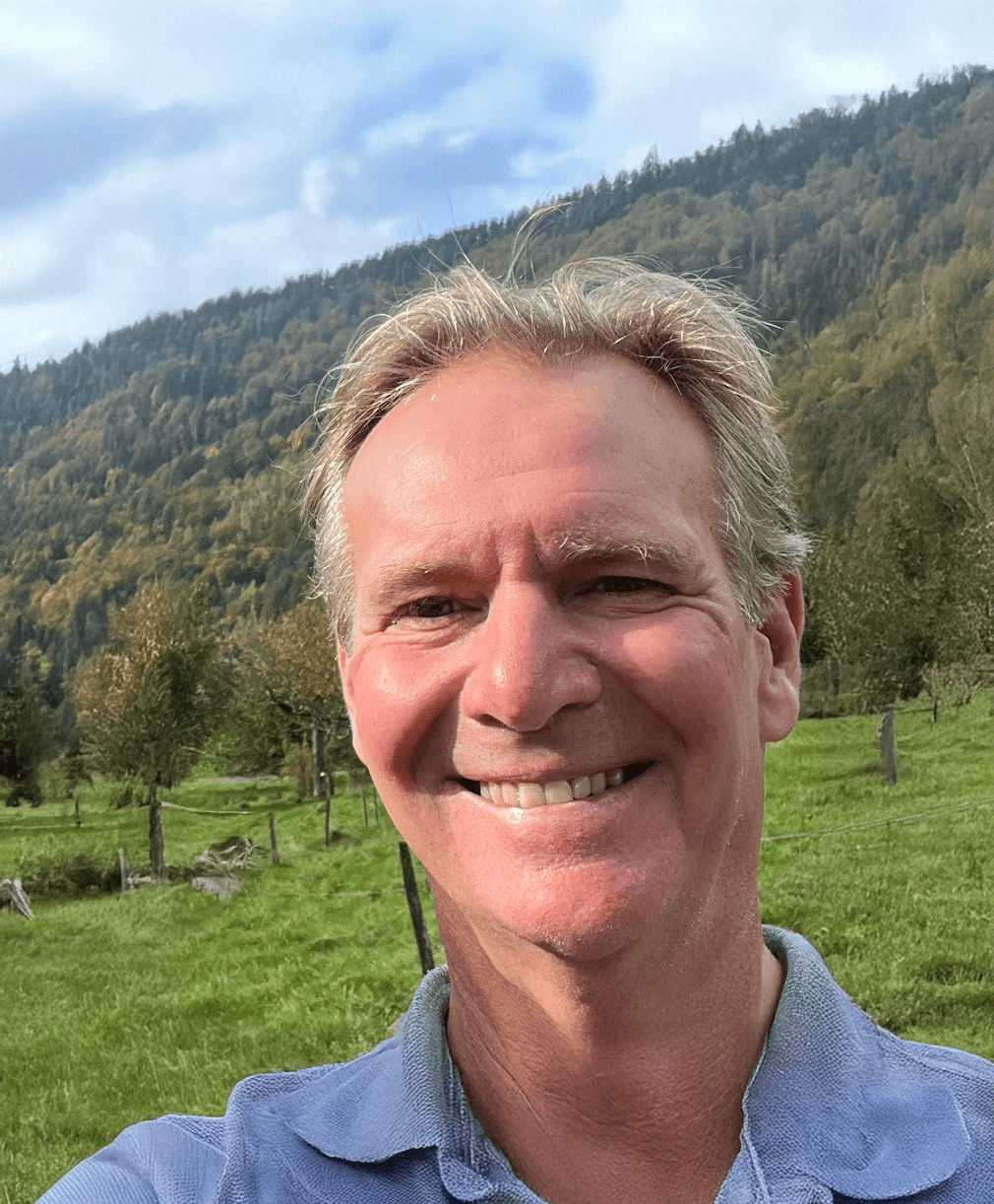 A smiling man taking a selfie in a green, hilly outdoor setting with trees and a mountain in the background.