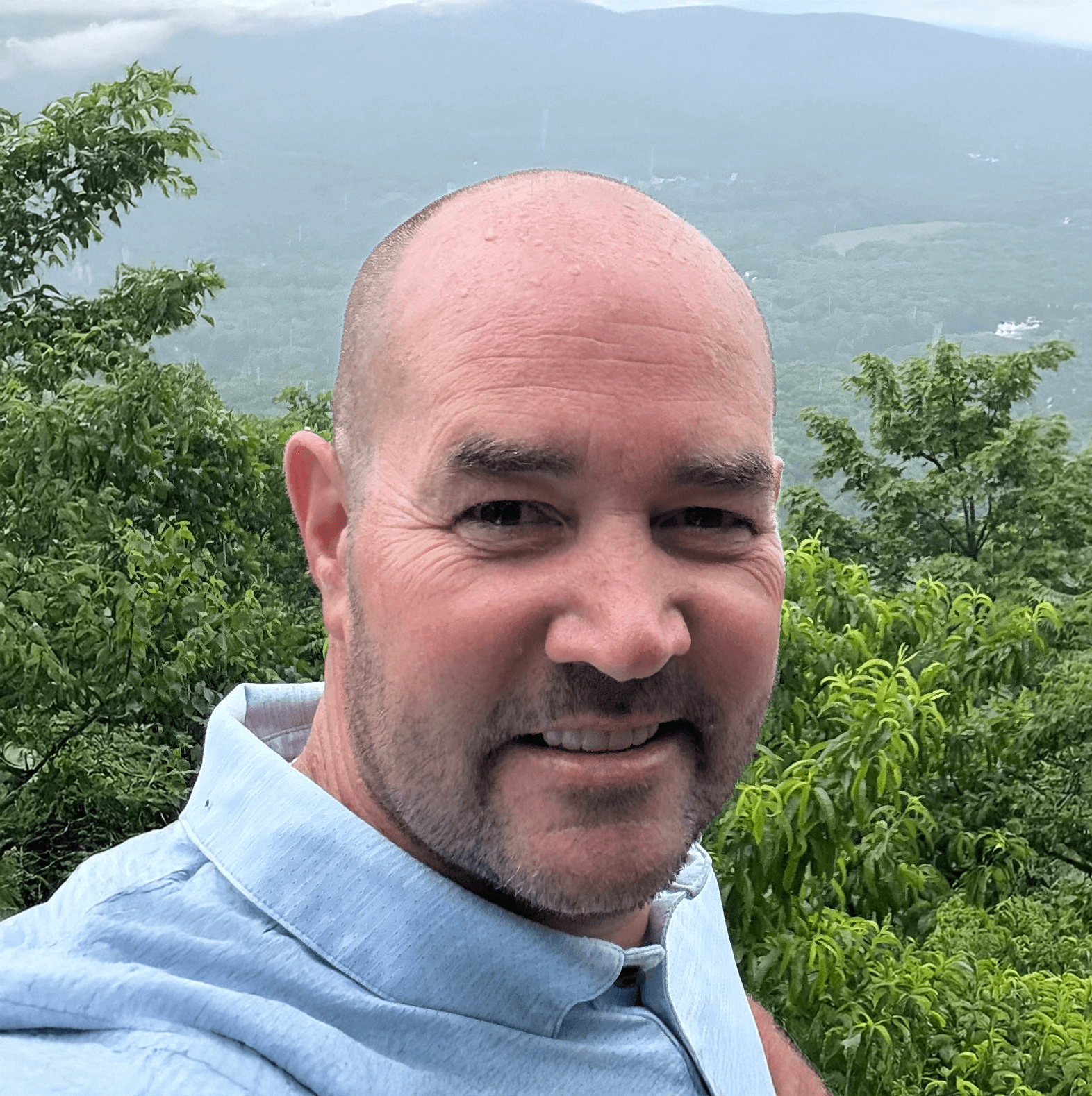 Close-up of a smiling man with a bald head and light facial hair, wearing a light blue shirt, outdoors with green trees and a foggy mountainous background.