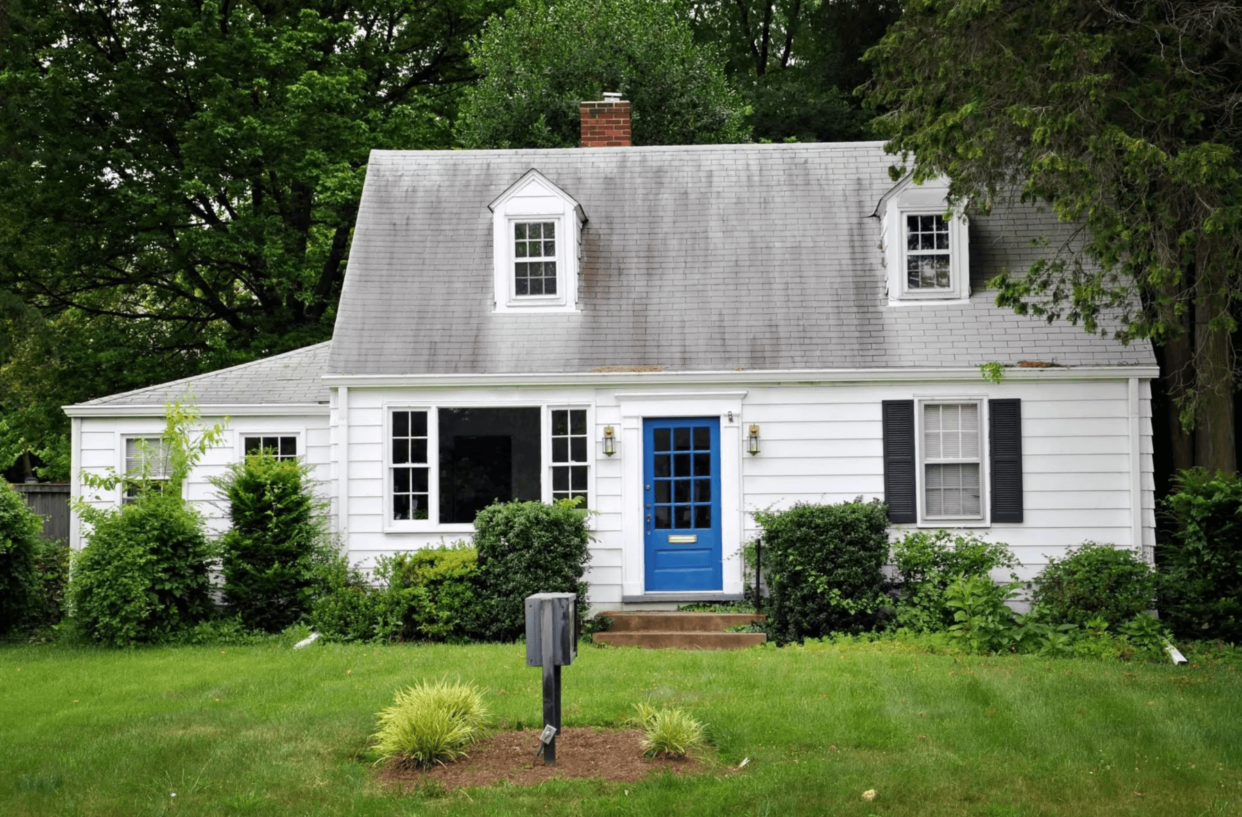 A white, two-story house with black shutters and a blue front door, surrounded by green bushes and trees, with a well-maintained lawn and a mailbox in the front yard.
