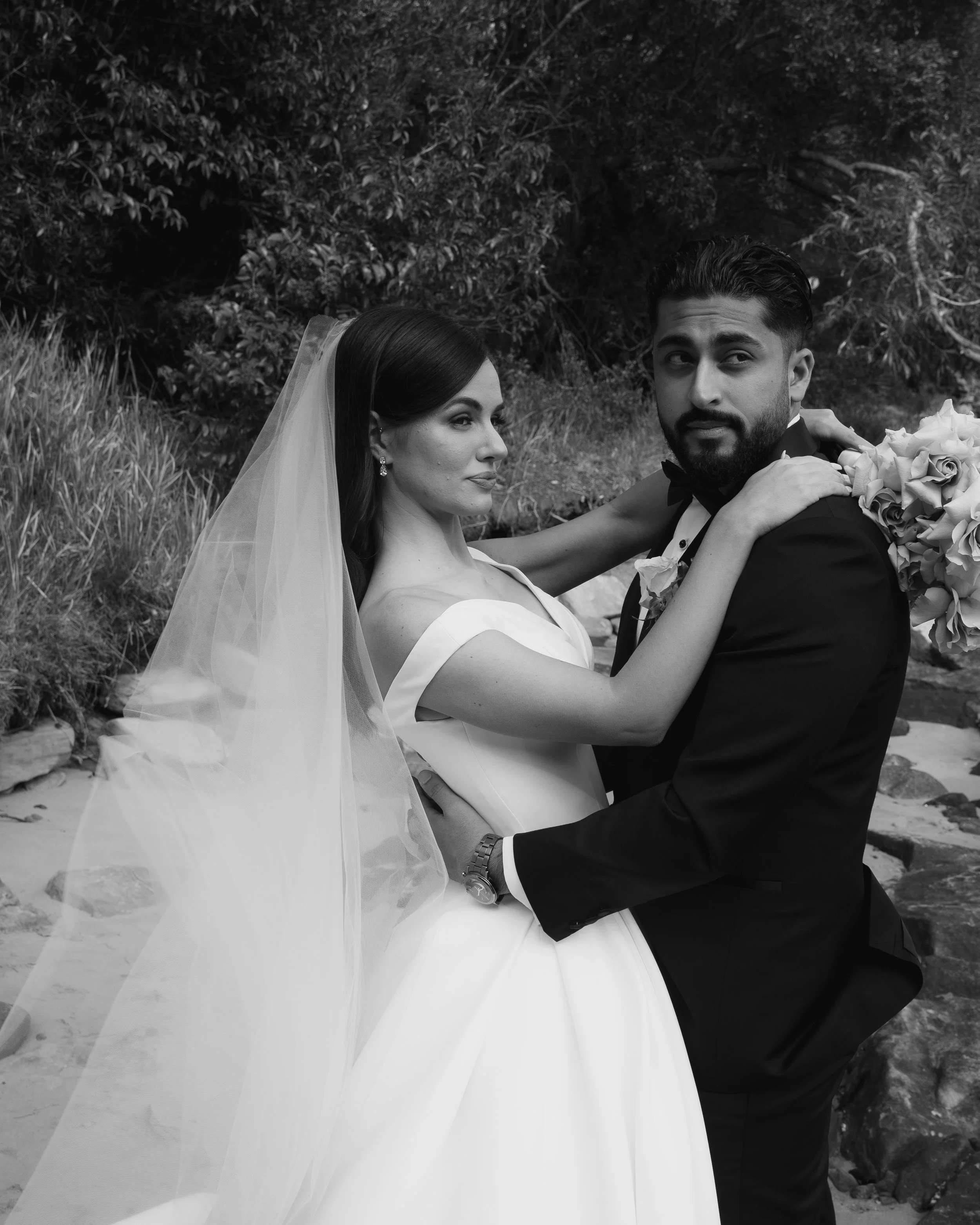 A bride and groom in wedding attire embrace outdoors, with the bride in a white dress and veil, and the groom in a tuxedo, holding a bouquet of flowers.