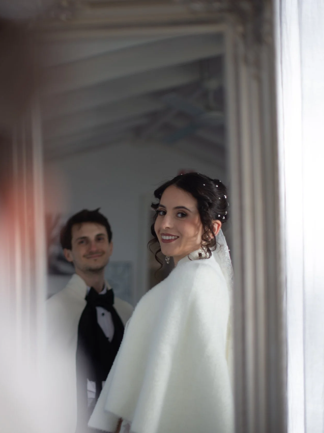 Bride smiling in a white dress and veil, looking into a mirror, with a young man in a tuxedo in the background.