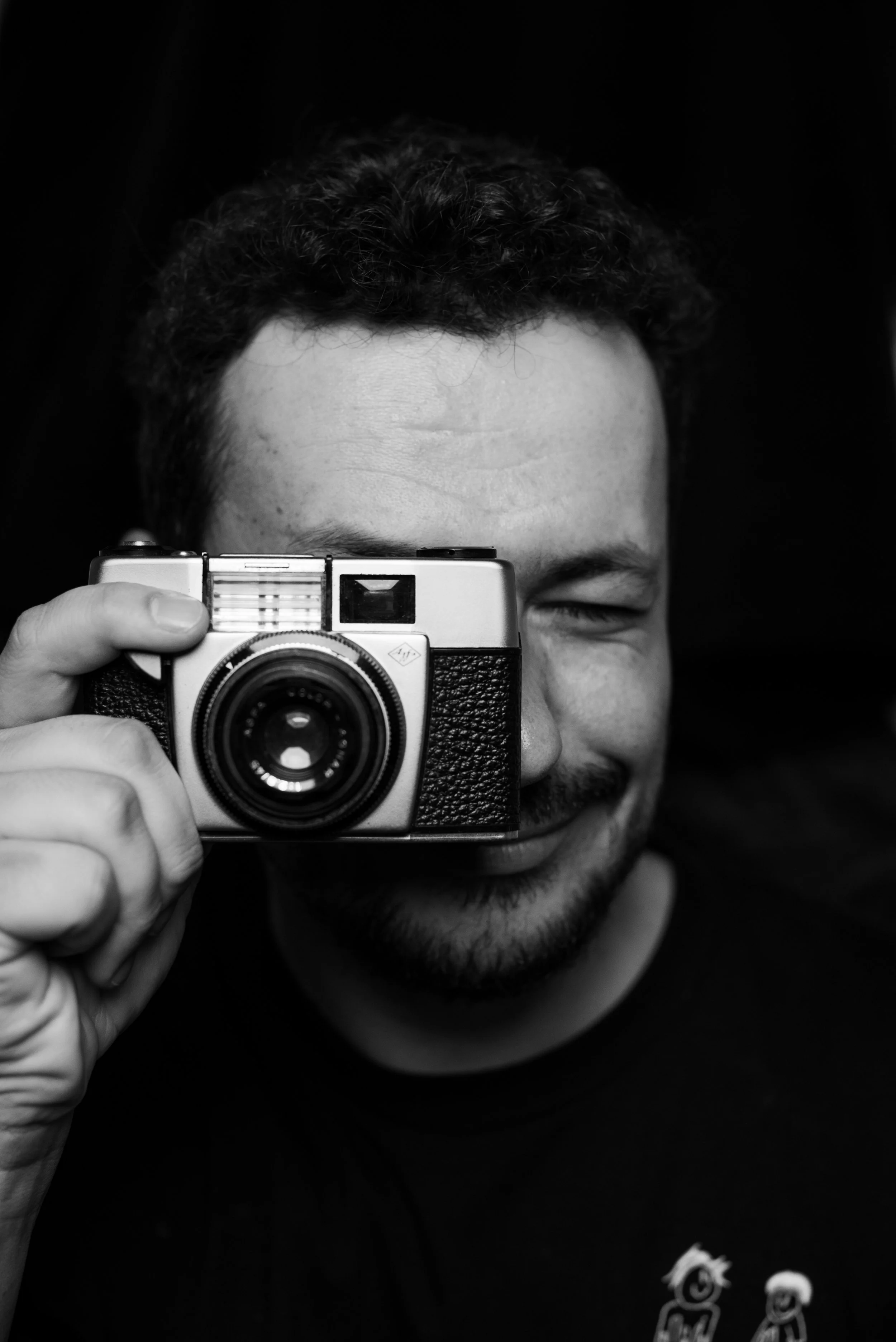 A smiling man with curly hair holding a vintage camera up to his face, partly covering his eye, with his other eye closed.