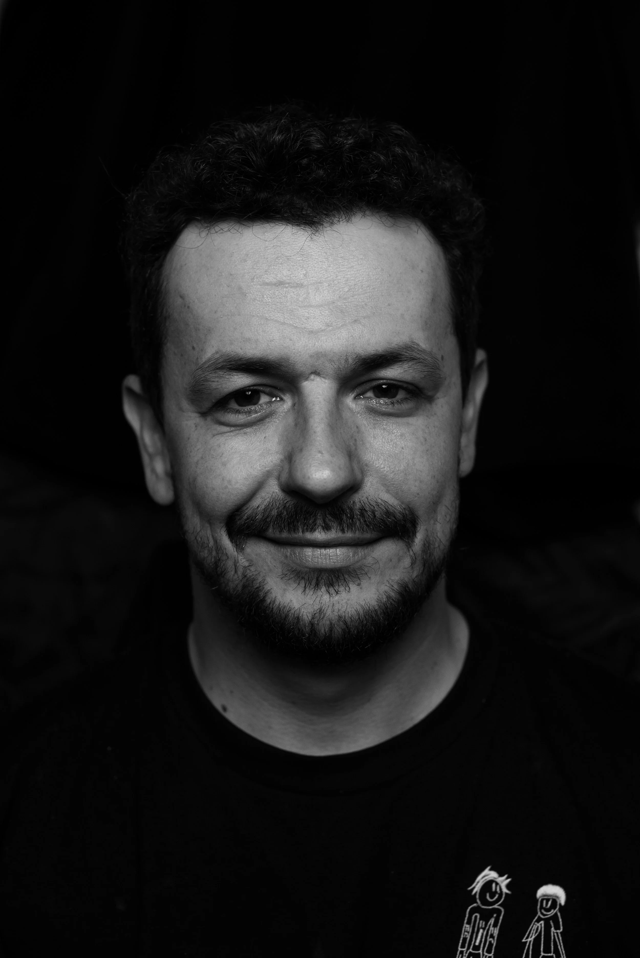 Black and white portrait of a smiling man with short curly hair, facial hair, and wearing a t-shirt with cartoon graphics.