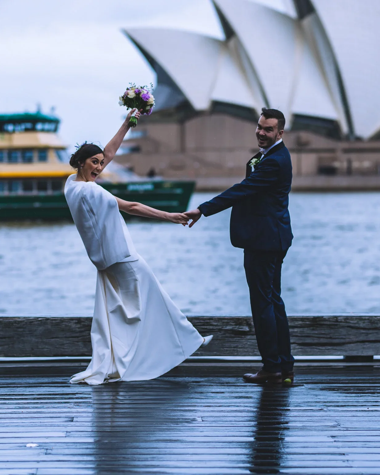 A bride and groom holding hands near the Sydney Opera House and water with a boat in the background, celebrating their wedding.