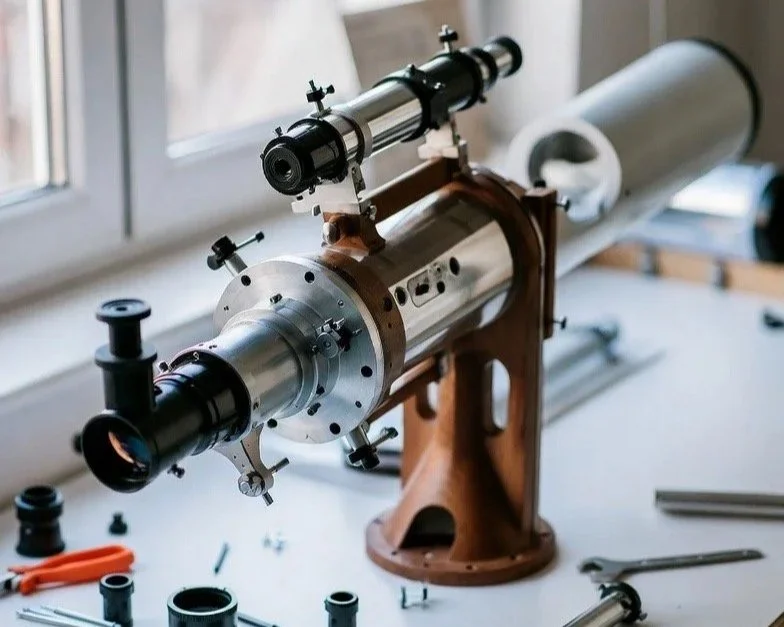 A telescope on a wooden mount set up on a white table near a window, with various tools and parts around it.