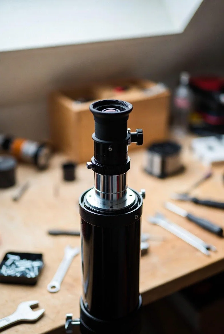 A telescope on a wooden workbench with various tools and small containers in the background.