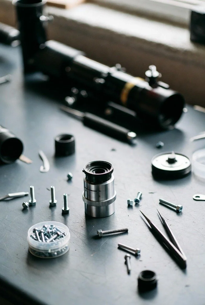 Disassembled telescope or camera lens with screws, bolts, and tweezers on a workbench near a black telescope.