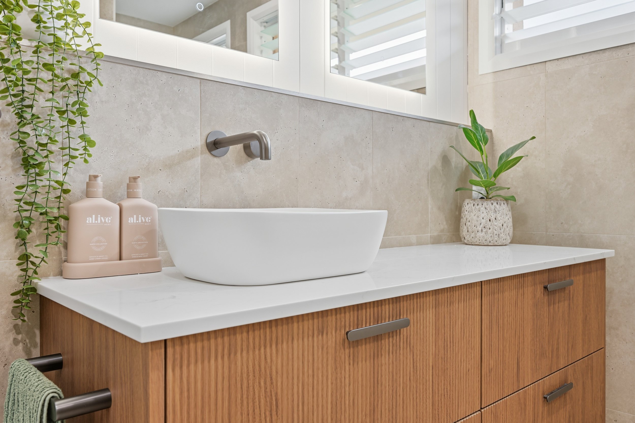 Bathroom vanity with a white vessel sink, two pink bottles of soap labeled 'alive', a green hanging plant, and a potted plant on a white countertop, beige tile wall, mirror, and a window with white blinds in the background.