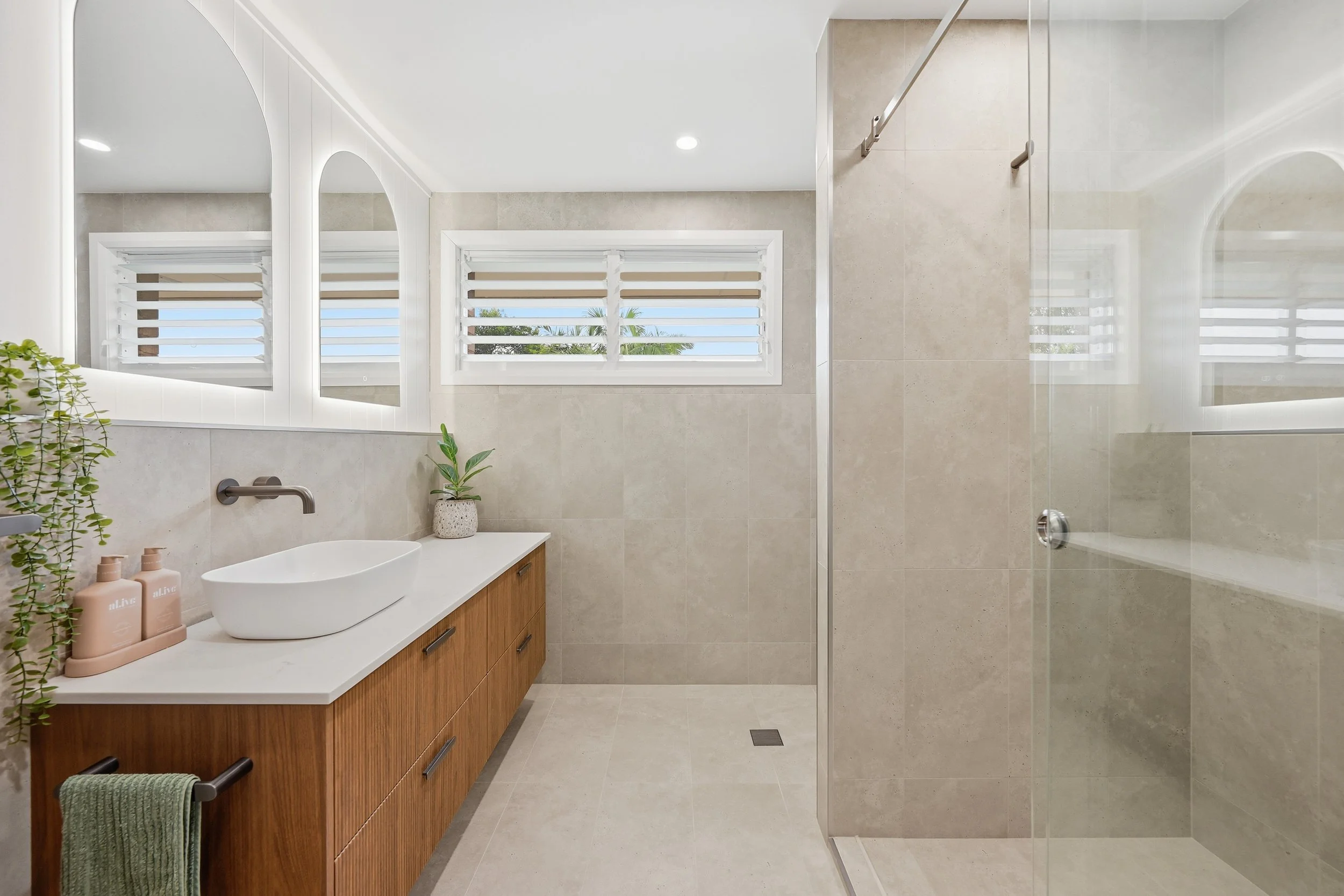 bathroom with tile walls and floor, a wooden vanity with a white vessel sink, two pink soap bottles, and a potted plant. There are two windows with horizontal blinds and a glass shower enclosure. Coffs Harbour Photography, Interior videography