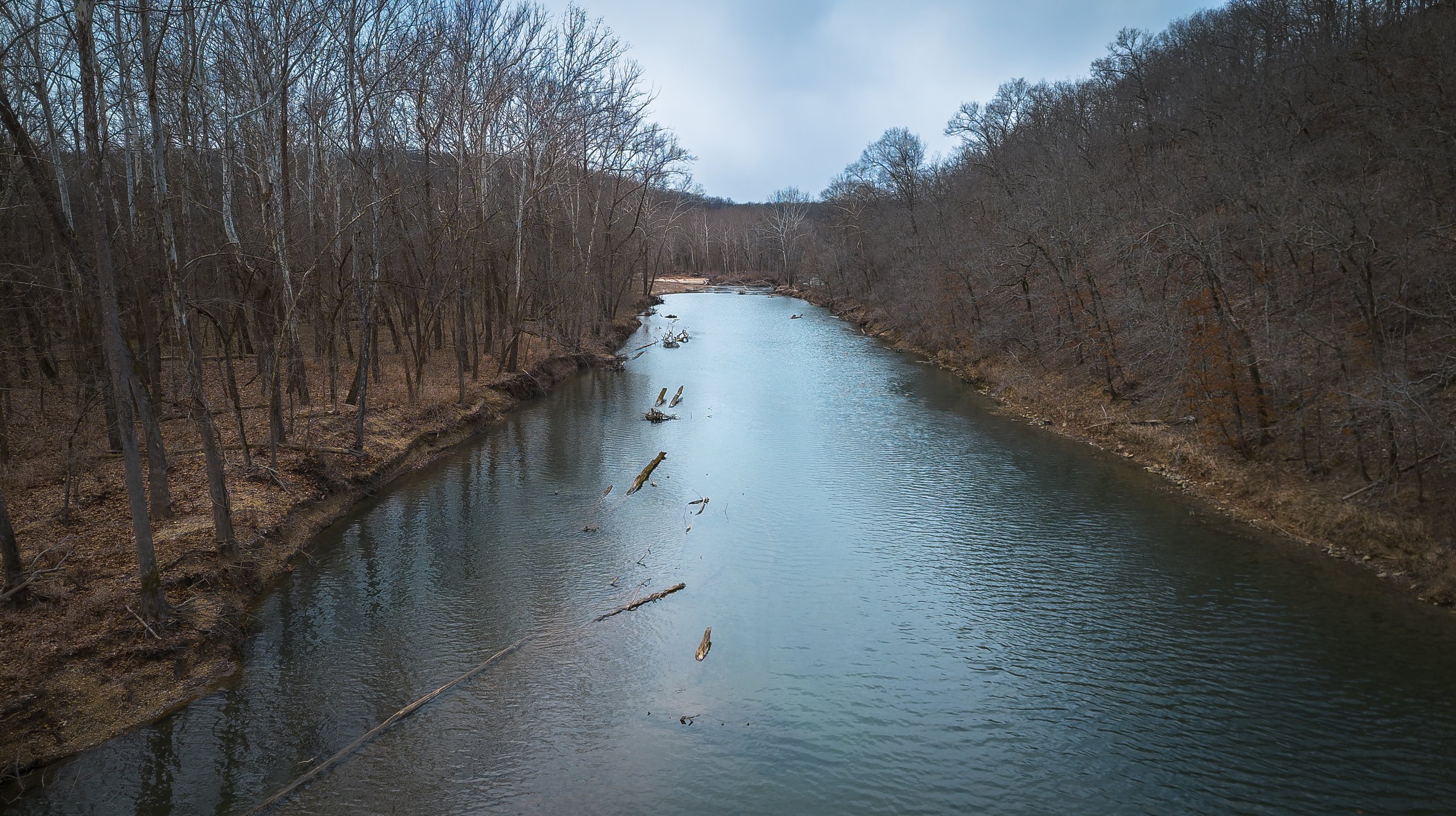 A river flowing through a wooded area with leafless trees on either side, some fallen branches in the water, and a cloudy sky above.