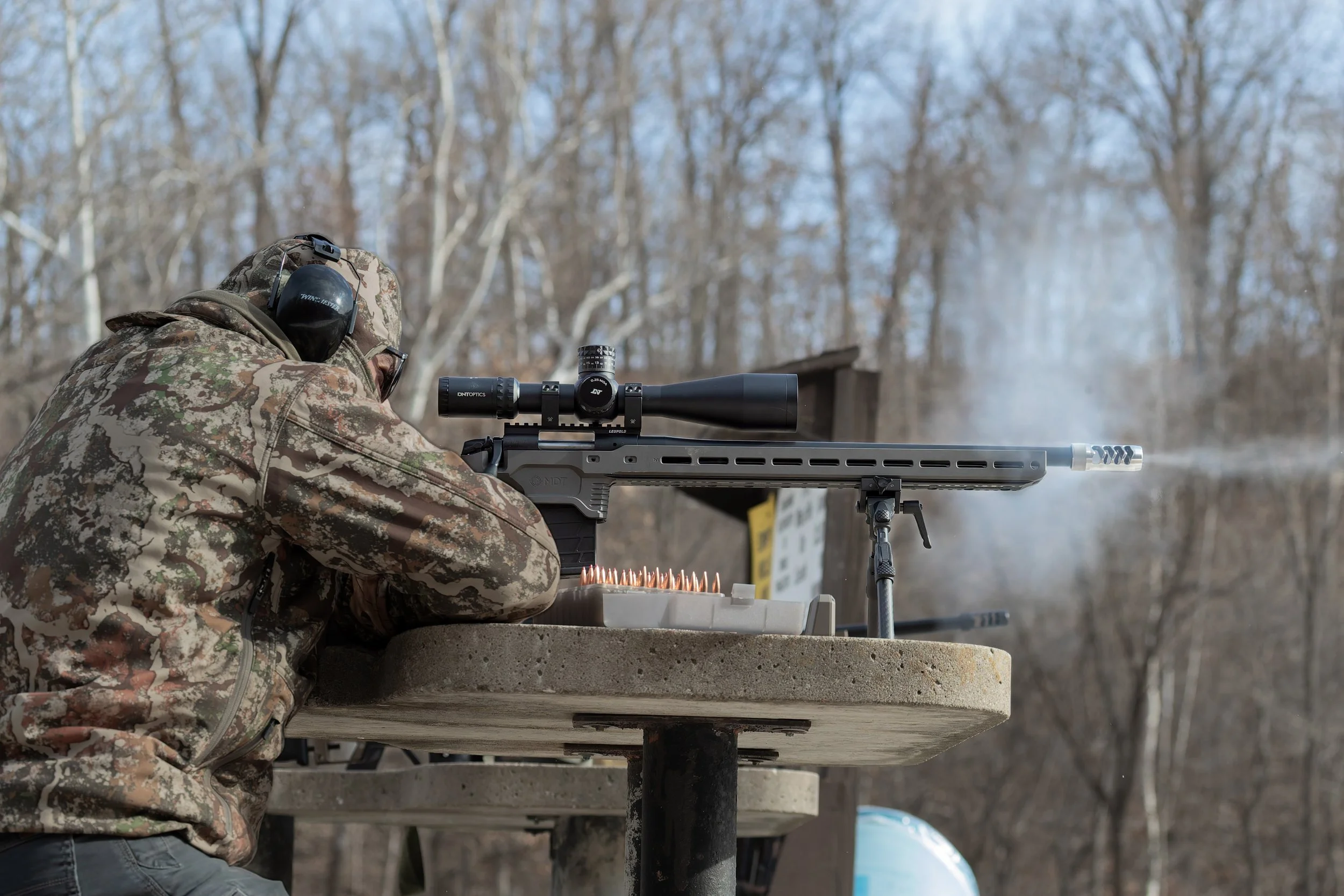 A person in camouflage clothing, wearing hearing protection, lying on a concrete surface, aiming a large sniper rifle with a scope and suppressor, outdoors in a wooded area.