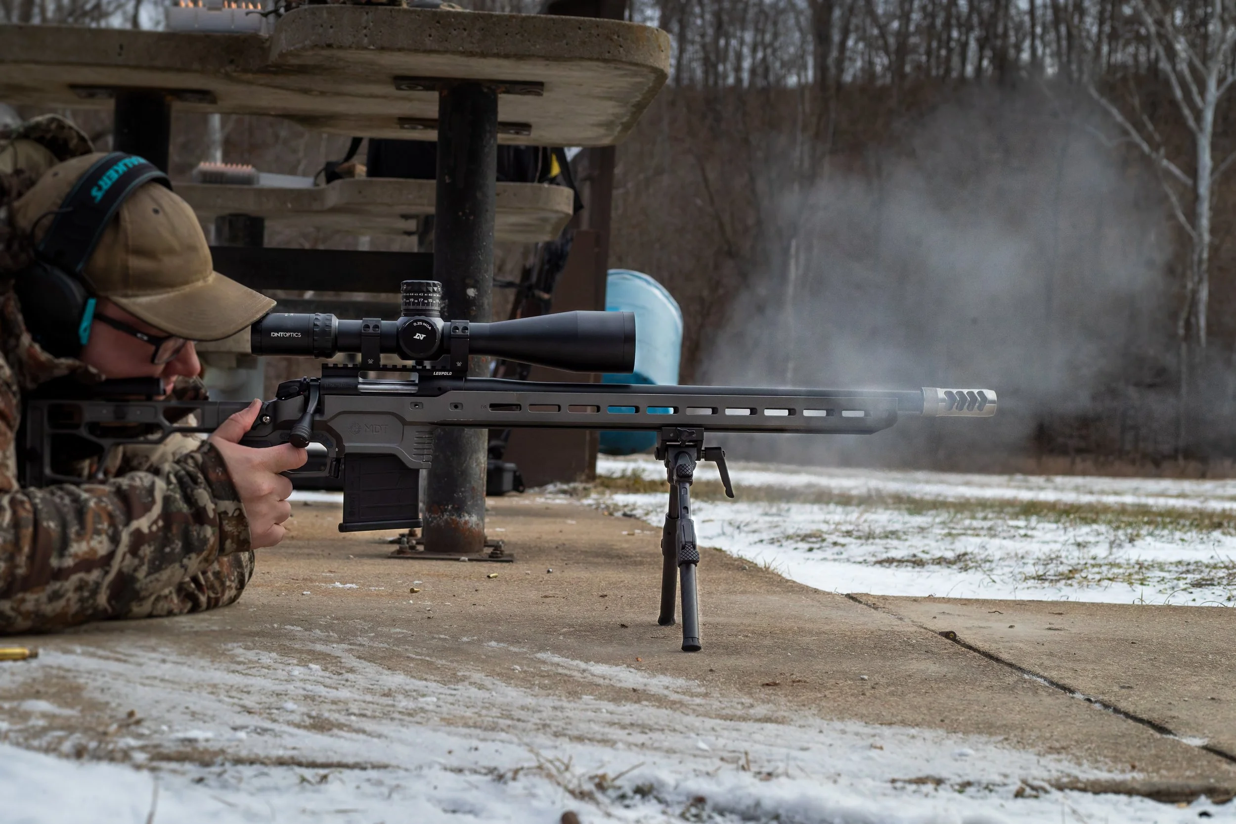 Person in camouflage clothing and tan cap lying prone on concrete, firing a large sniper rifle with a scope, outdoors in a winter landscape with snow on the ground and leafless trees in the background.