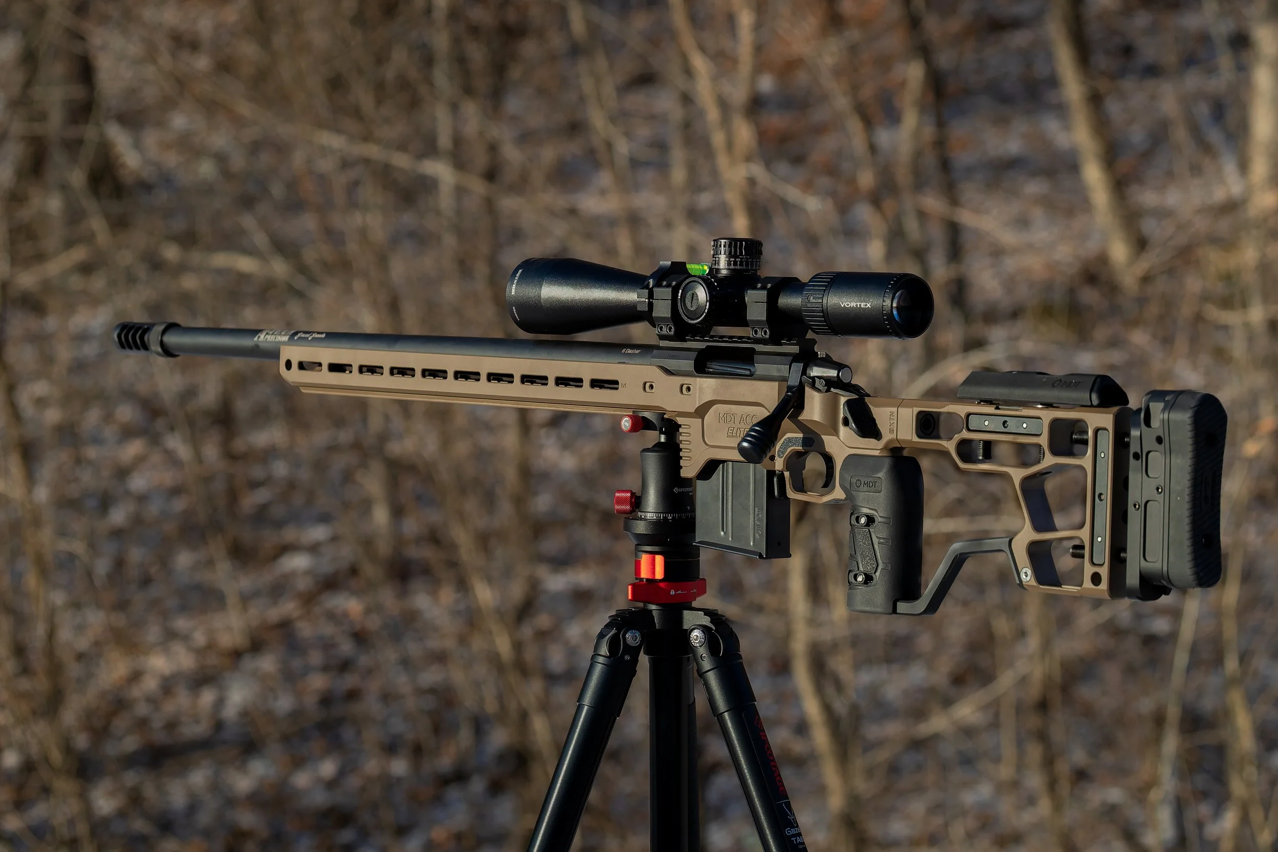 A tactical rifle with a scope mounted on a tripod, set outdoors in a wooded area.